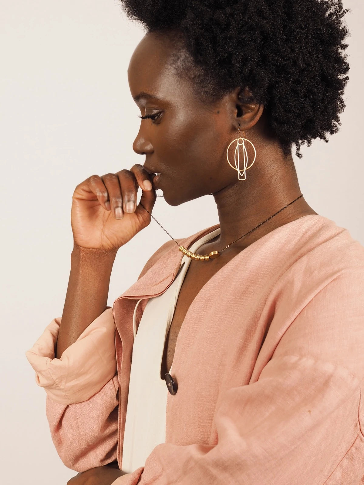 A woman with short curly black hair is shown in profile wearing large gold geometric hoop earrings, a Nucleo Solid Brass Totems and Brass Disc Necklace by brass+bold, and a dusty pink linen shirt over a white top, set against a plain light beige background.