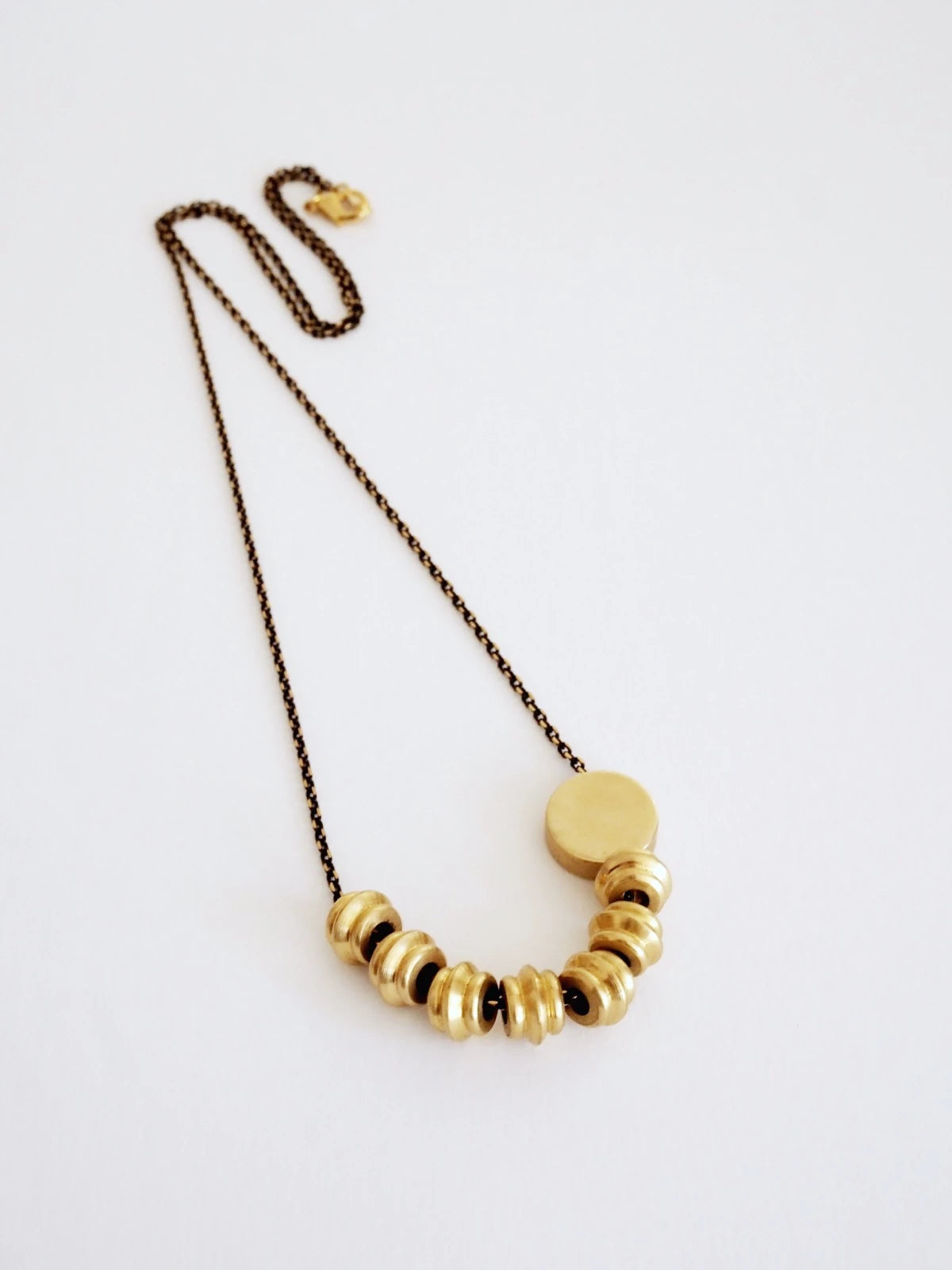 Nucleo Solid Brass Totems and Brass Disc Necklace by brass+bold featuring a curved row of textured brass discs on a dark bronze chain, displayed on a clean white background.