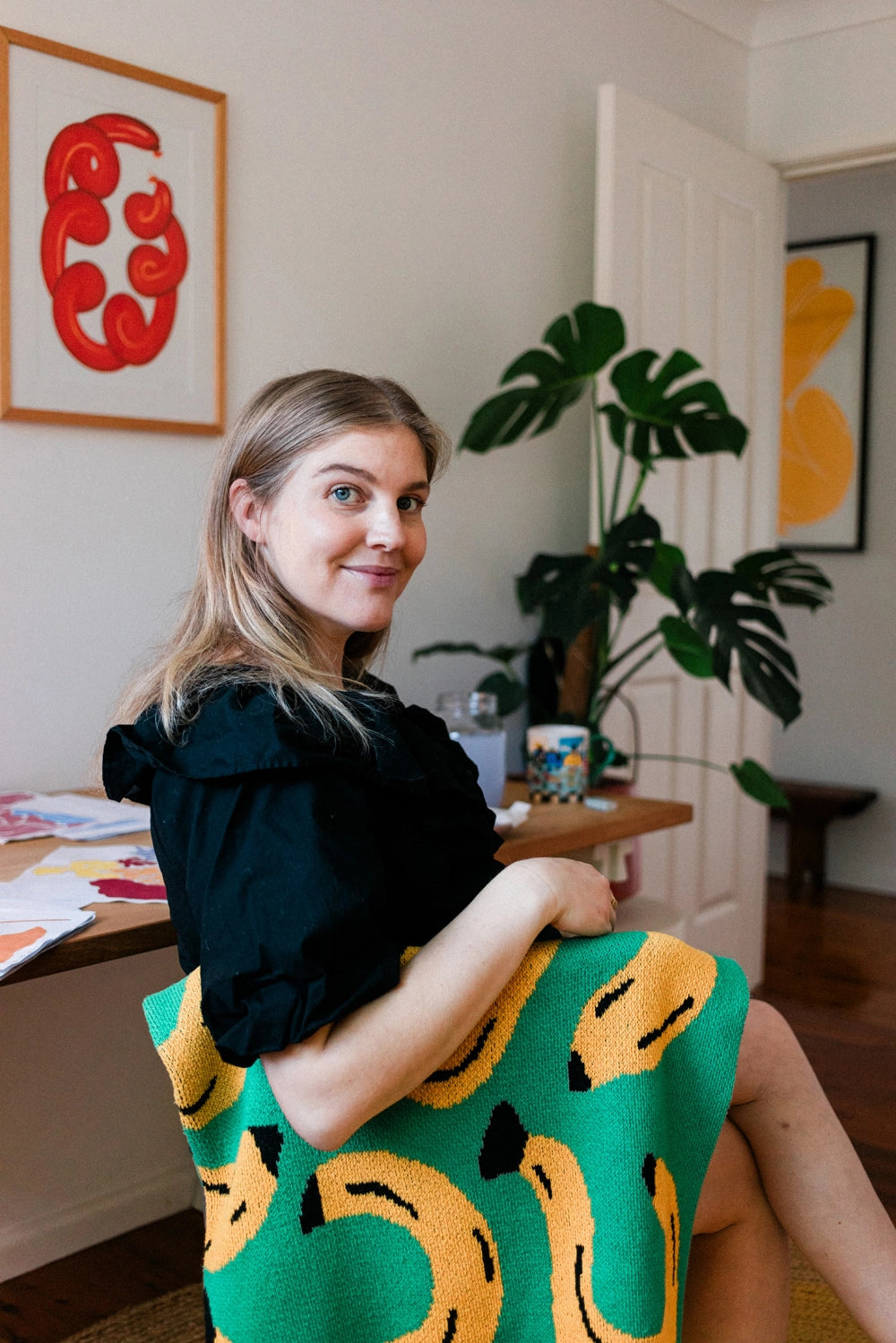 Illustrator Liv Lee in her studio sitting on a chair covered in a bright green throw with bananas on it