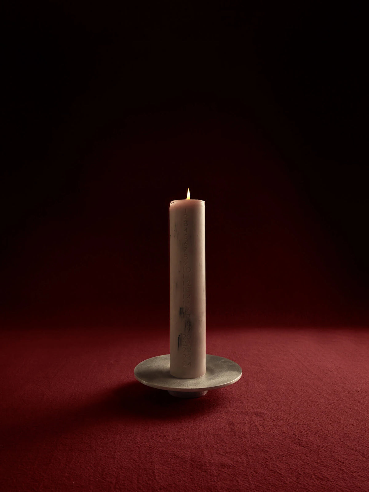 A single lit taper Mura advent candle by ferm living in Orchid colour in a holder, photographed against a dark red and black studio background.