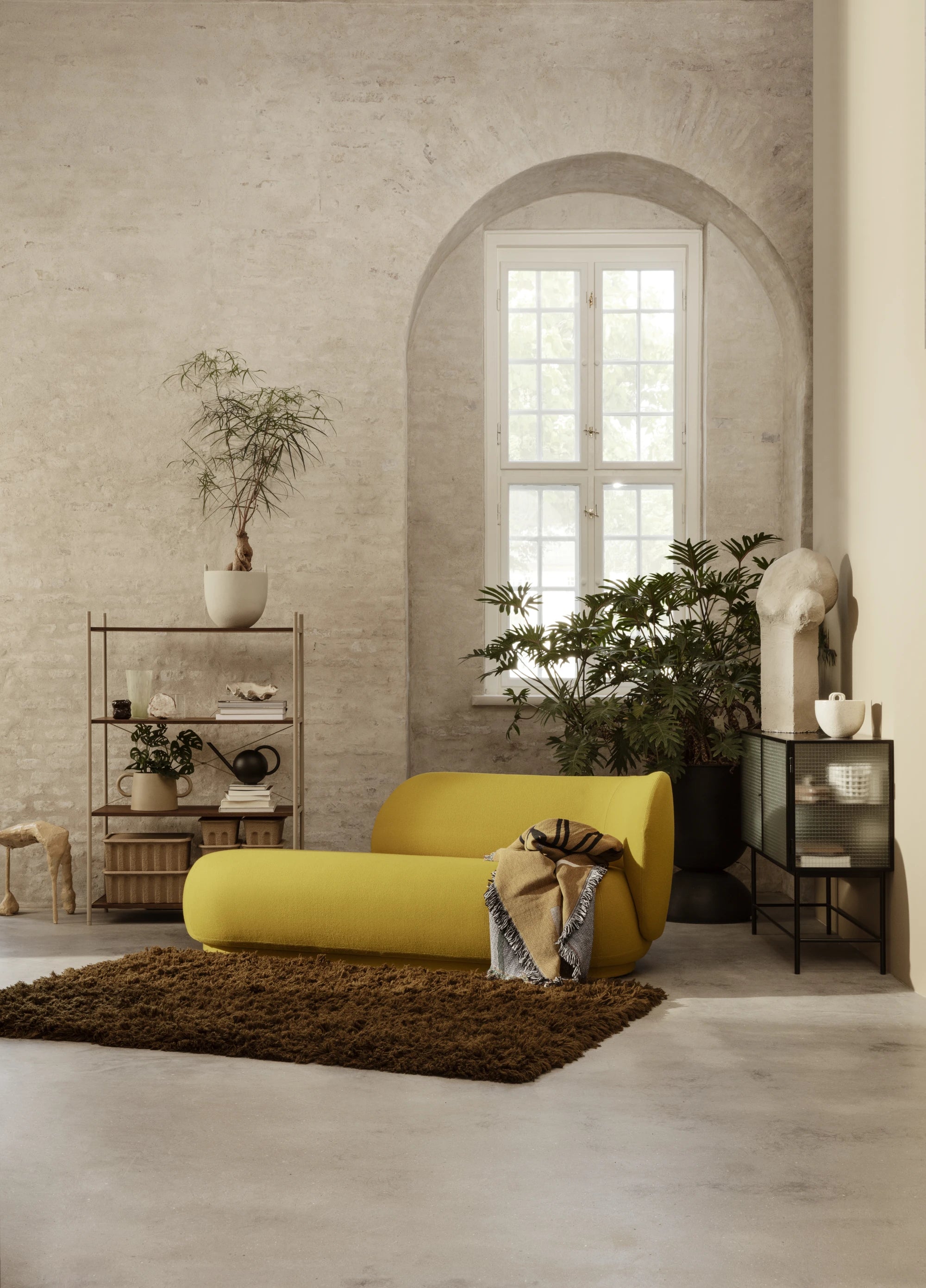 Ferm Living Rico Divan in gold toned fabric with right sitting backrest in a bright stone walled room with a brown  high pile rug on the floor