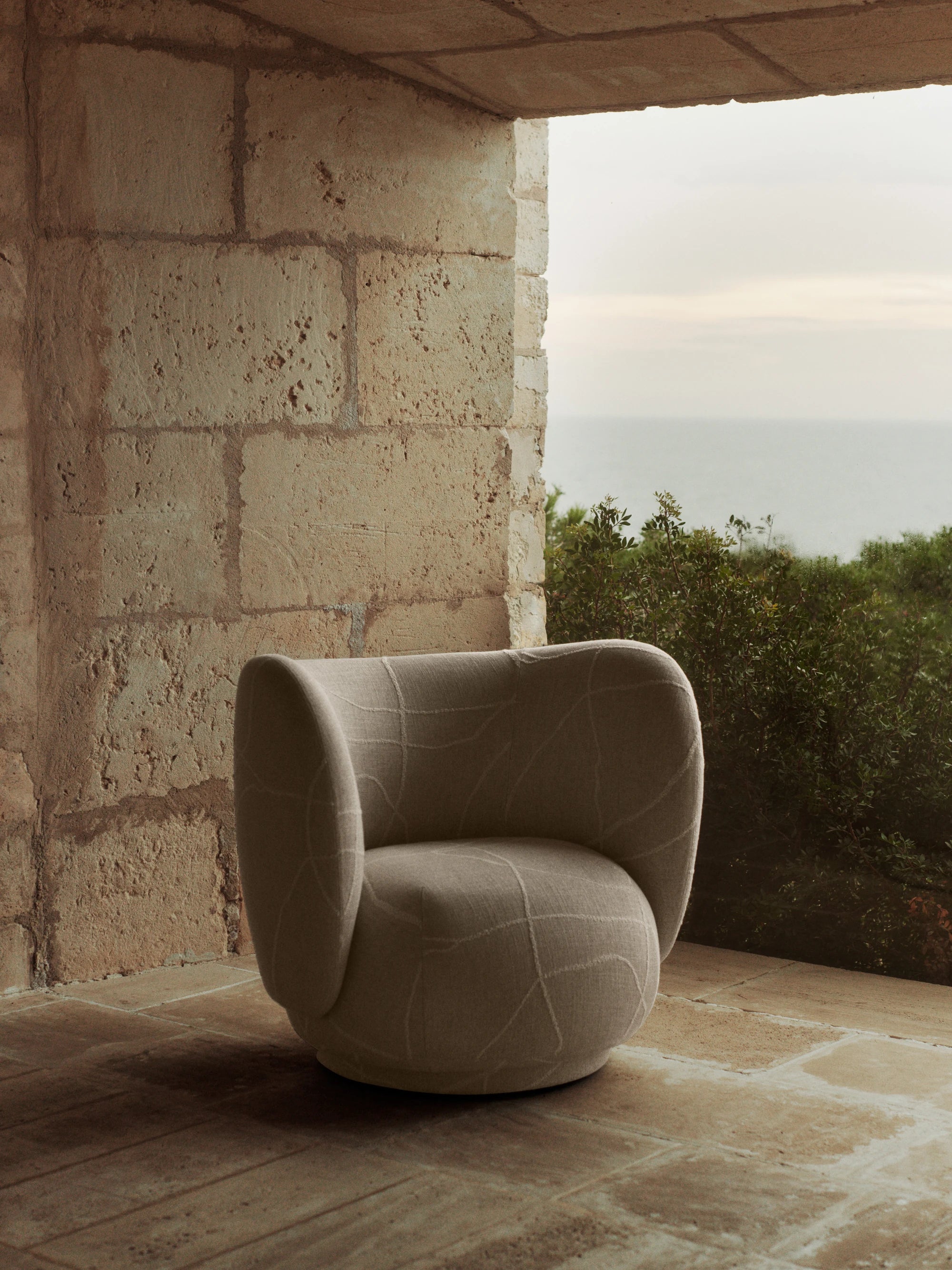 Ferm Living Rico Lounge Chair in Utzon-can lis fabric standing on a flagstone floor against a stone wall and open door frame to greenery