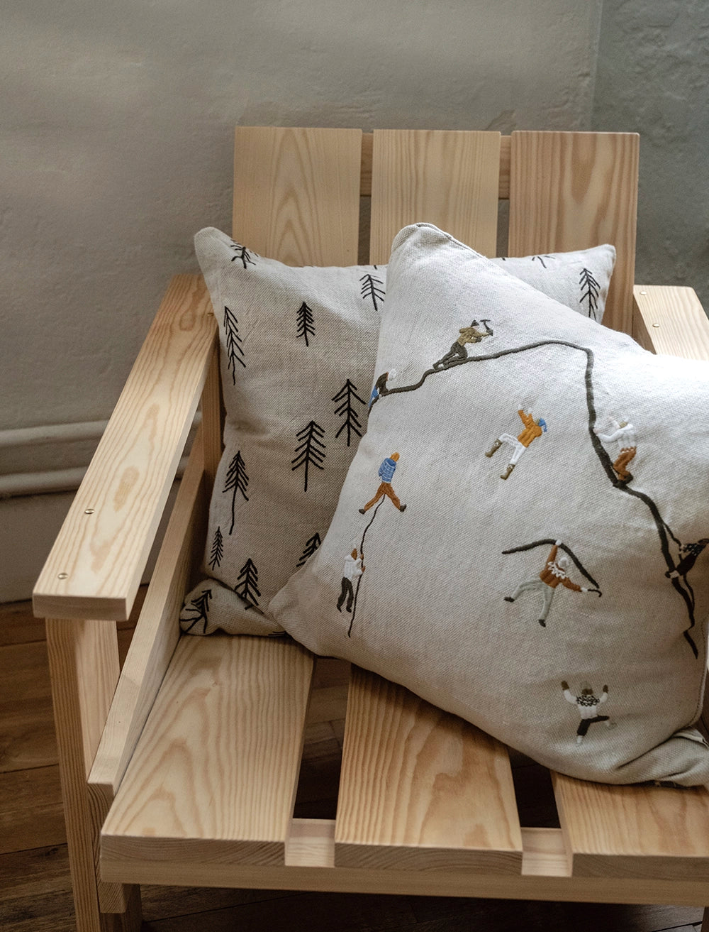 Fine Little Day 'Mountain Climbers' Embroidered Square Cushion placed on a wooden chair next to a natural cushion with embroidered trees