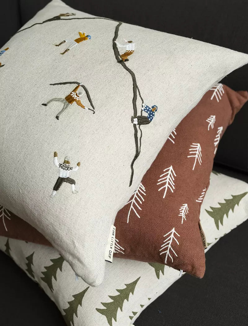 Fine Little Day 'Mountain Climbers' Embroidered Square Cushion sitting atop aa brown cushion with embroidered trees and a natural cushion with green trees