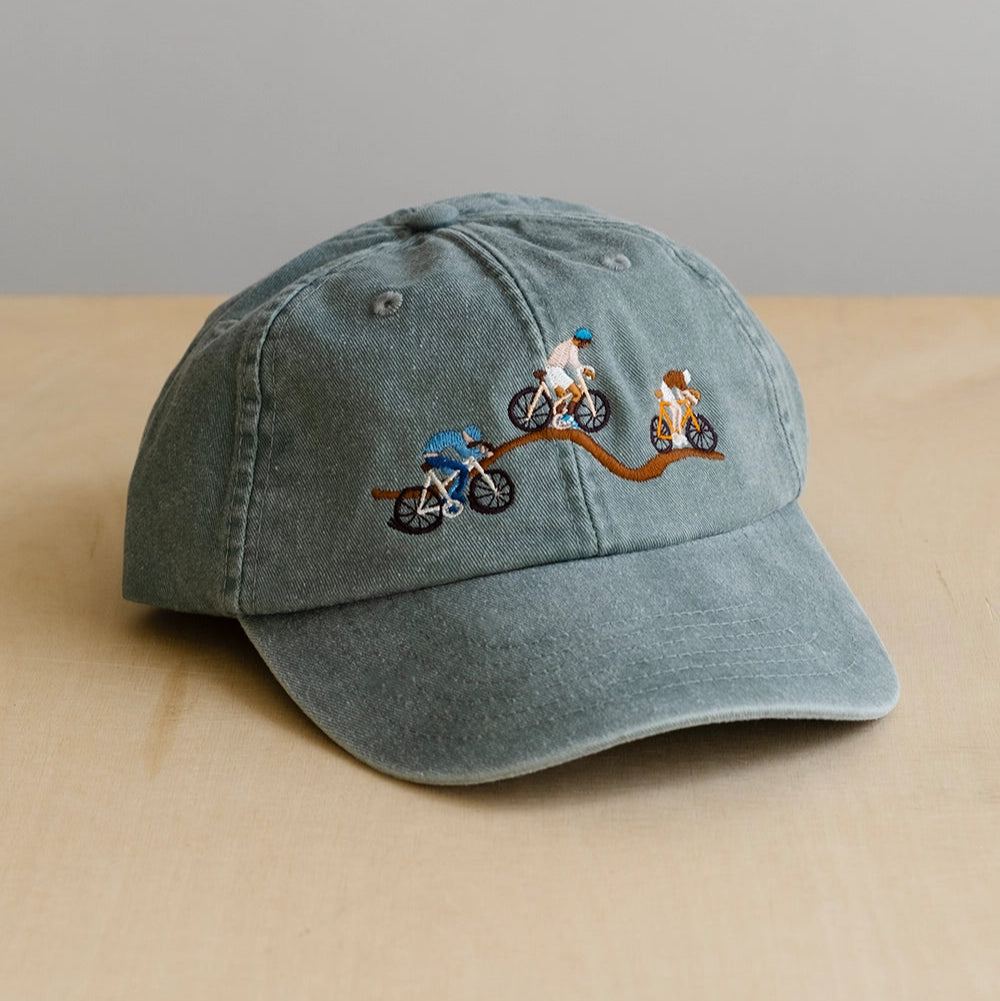 Washed green 6 panel baseball cap with embroidered designs of bikers on a path on a beige surface