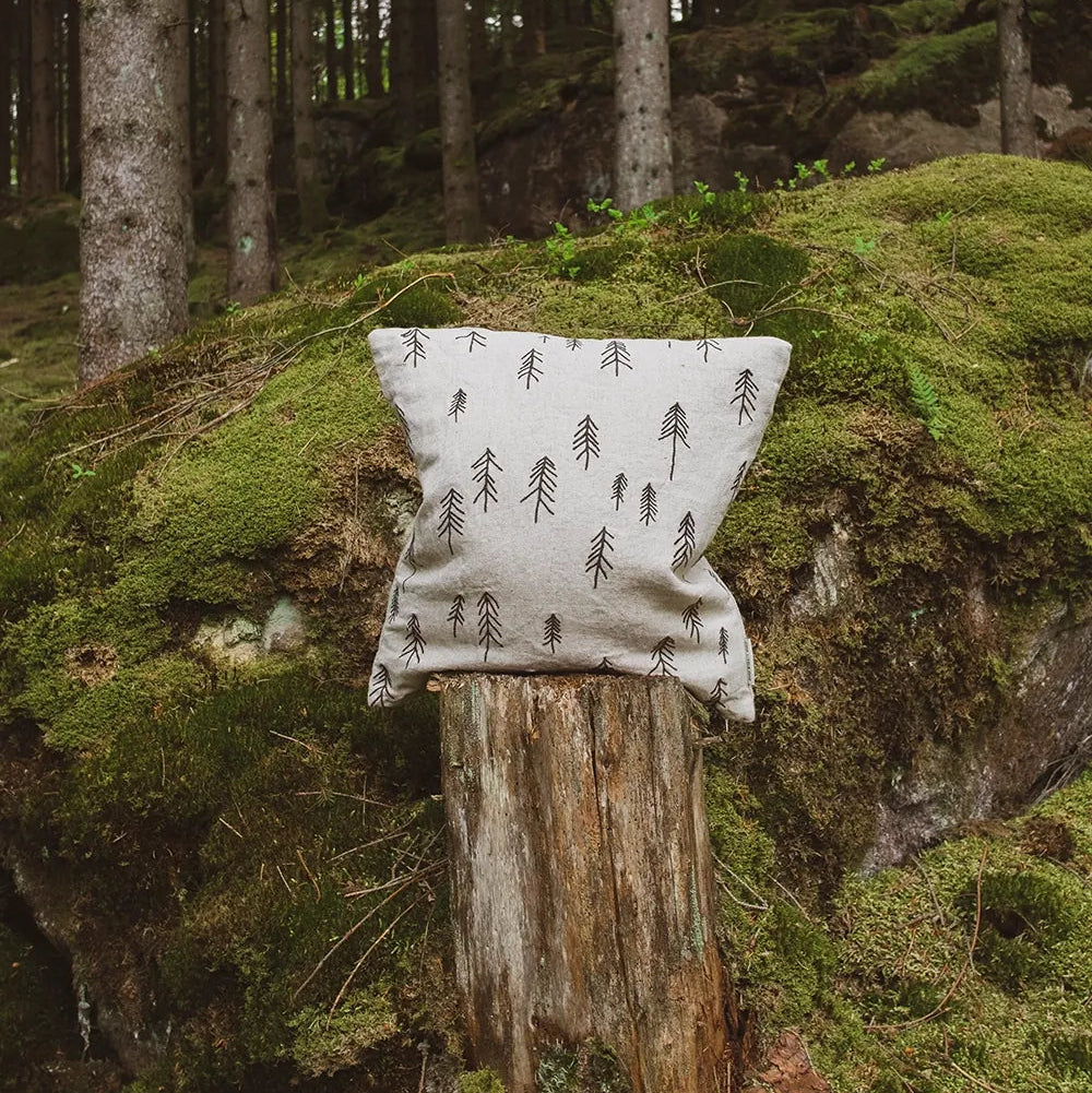 A square throw pillow with a black pine tree pattern on a light beige linen-like fabric, resting on a weathered tree stump in a mossy forest setting.