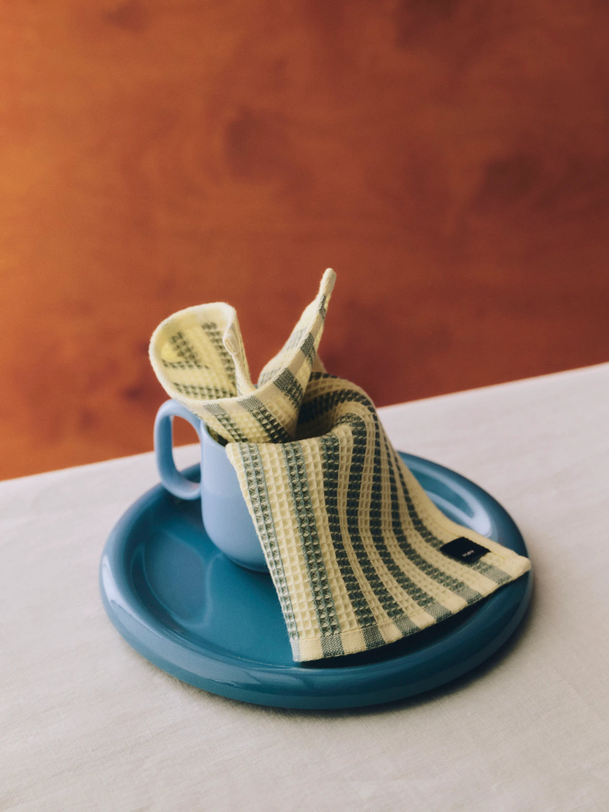 A folded HAY Canteen yelow and green checkered wash cloth is draped over a light blue ceramic mug, which sits on a round teal saucer. The scene is set on a light-colored wooden table against a textured, warm orange-brown background.