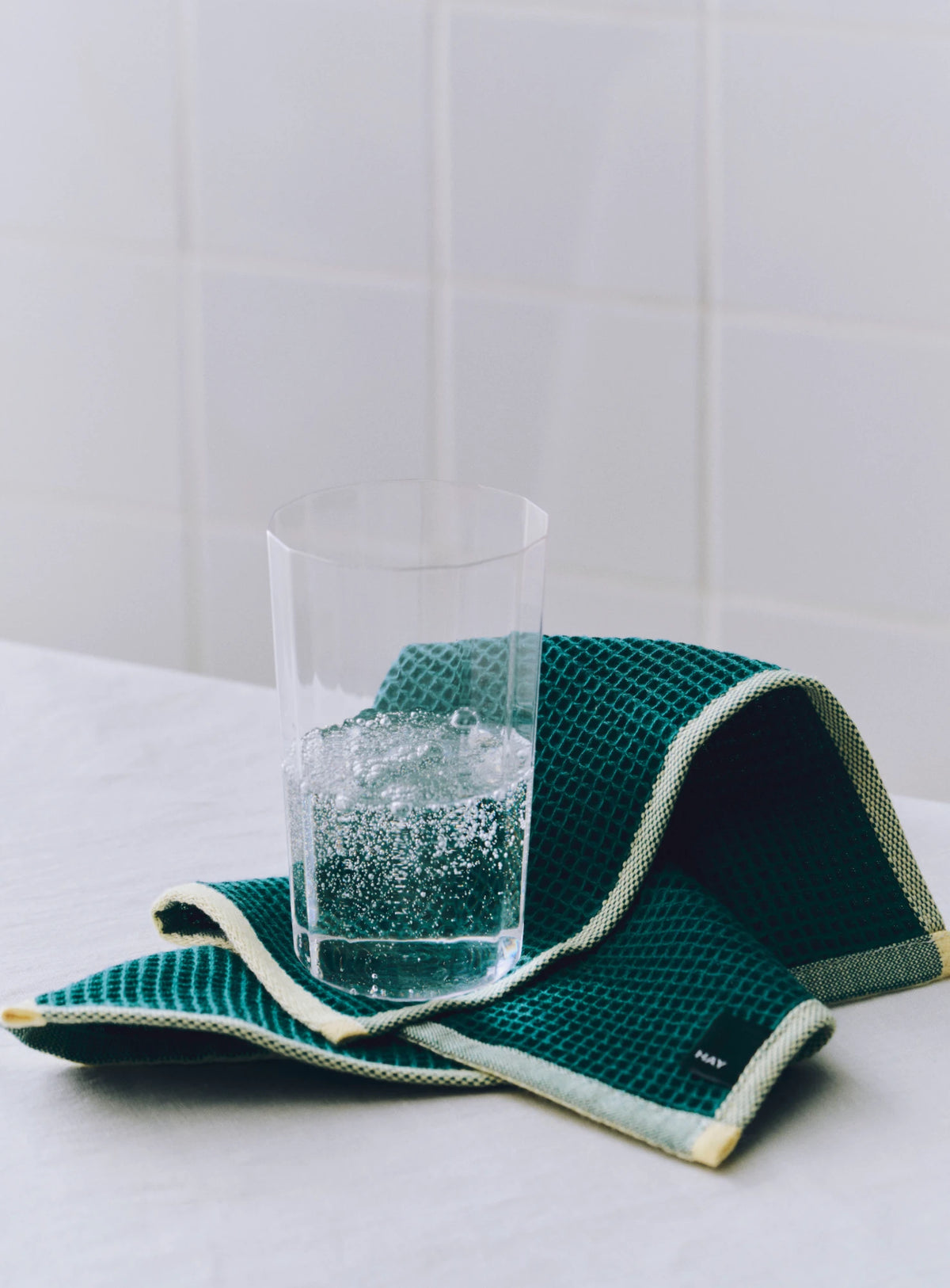 A dark green HAY Canteen waffle-weave wash cloth with cream trim, draped on a white tiled countertop with a clear glass of sparkling water resting on it.