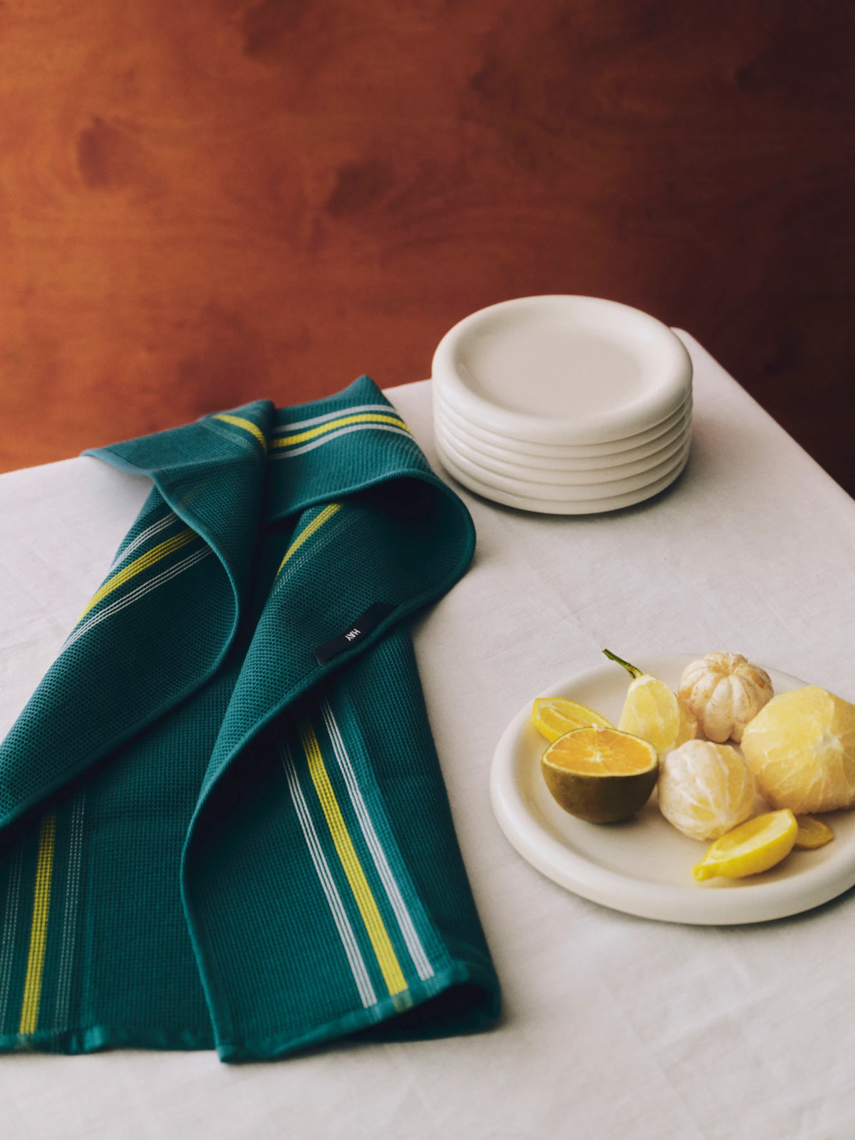 A folded dark teal HAY Canteen kitchen towel with yellow and white stripes is draped on a white tablecloth next to a stack of white plates and citrus fruit on a white plate, set against a warm brown background.