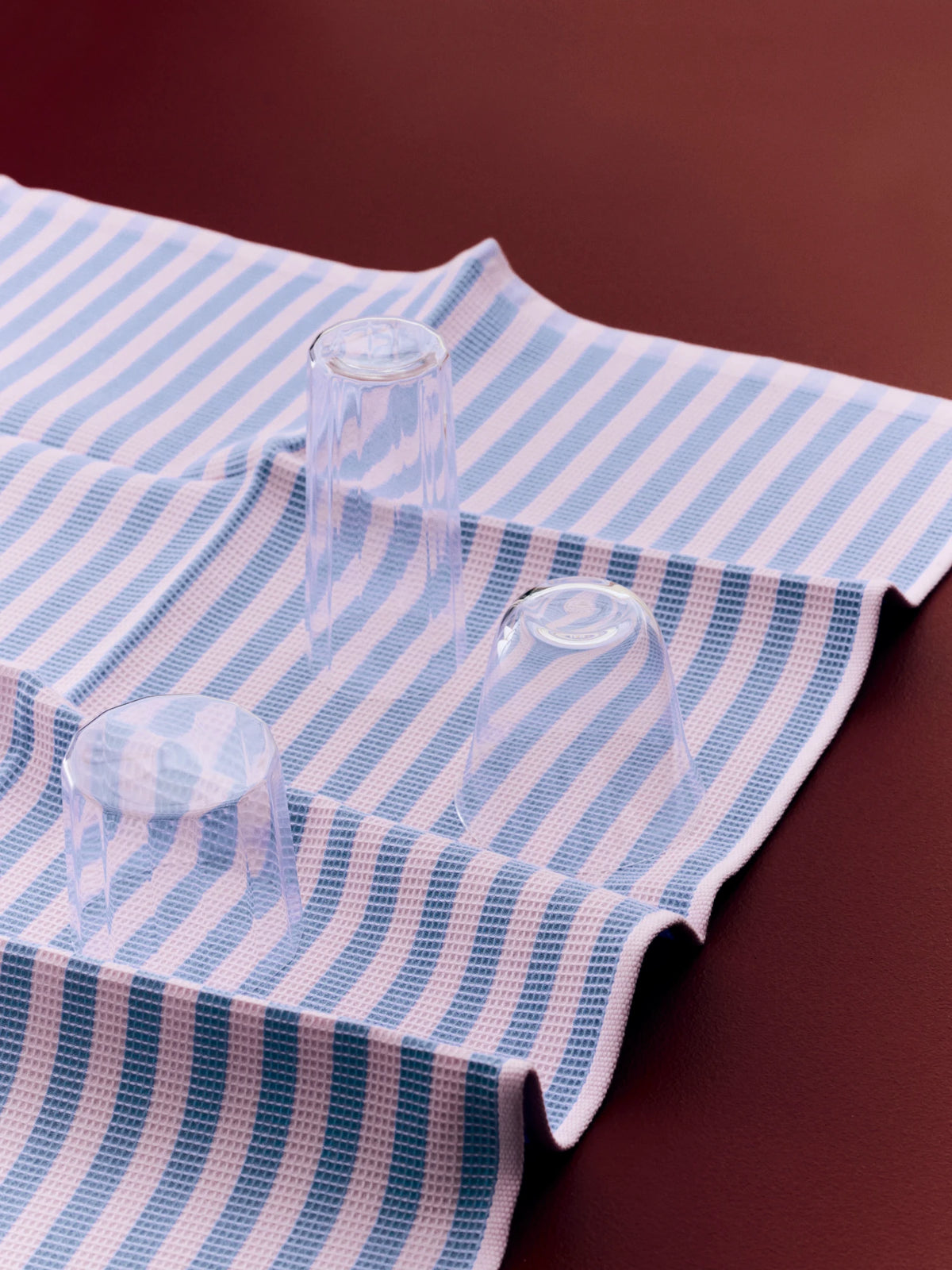 A close-up of a striped HAY Canteen kitchen towel or cloth with blue and pink vertical stripes, draped on a dark reddish-brown surface with three clear glass cups placed on top.