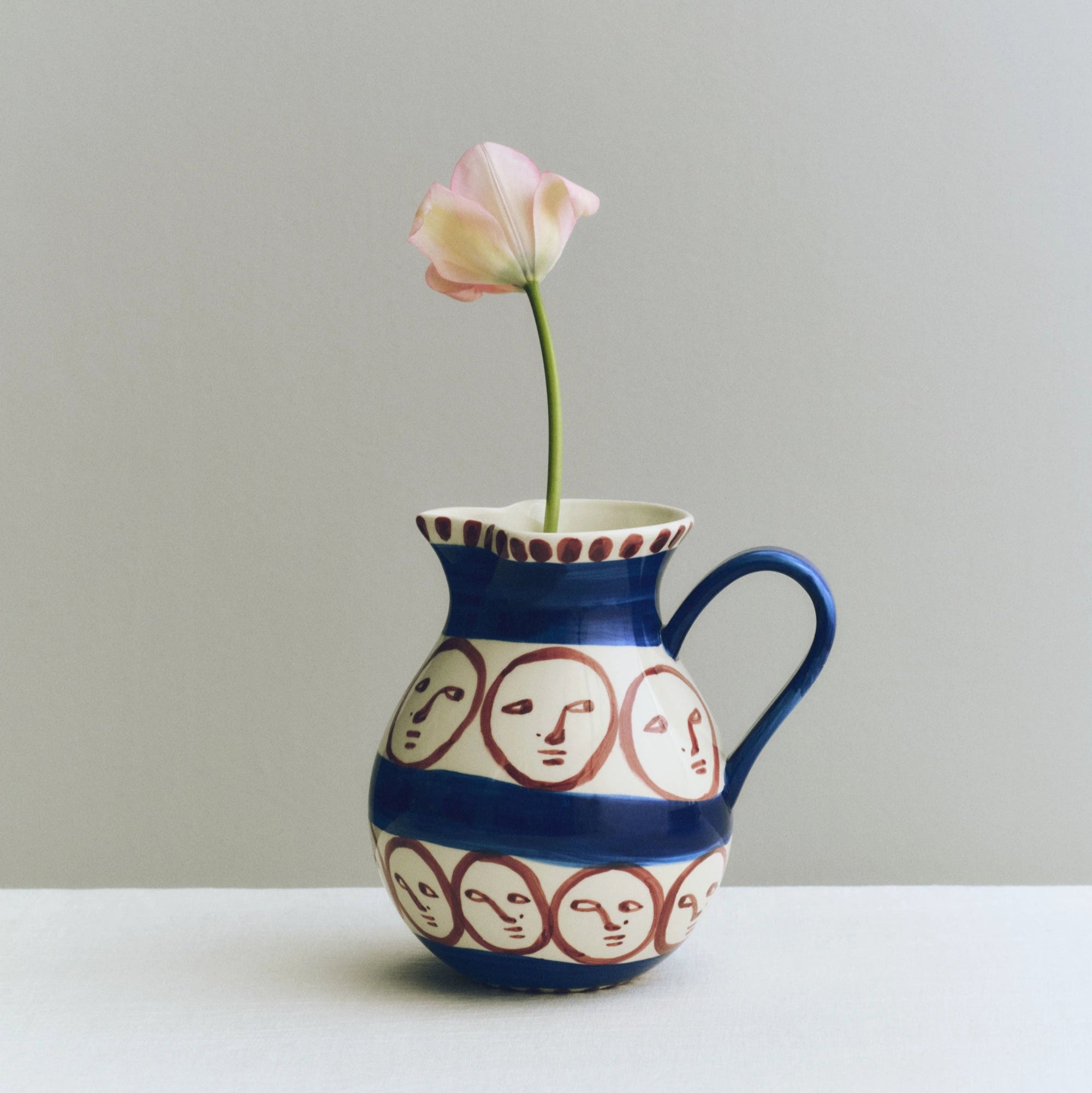 Ceramic pitcher with face patterns holding a single pink flower against a plain background