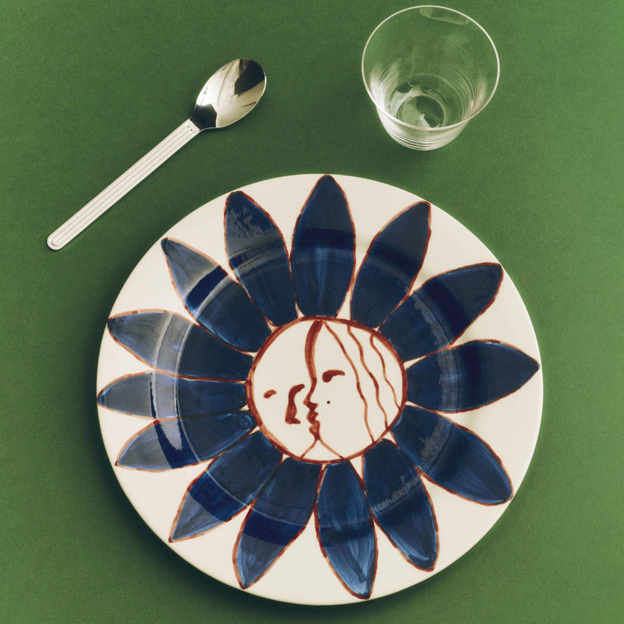 Ceramic plate with blue floral design and face on a green surface with a spoon and glass.
