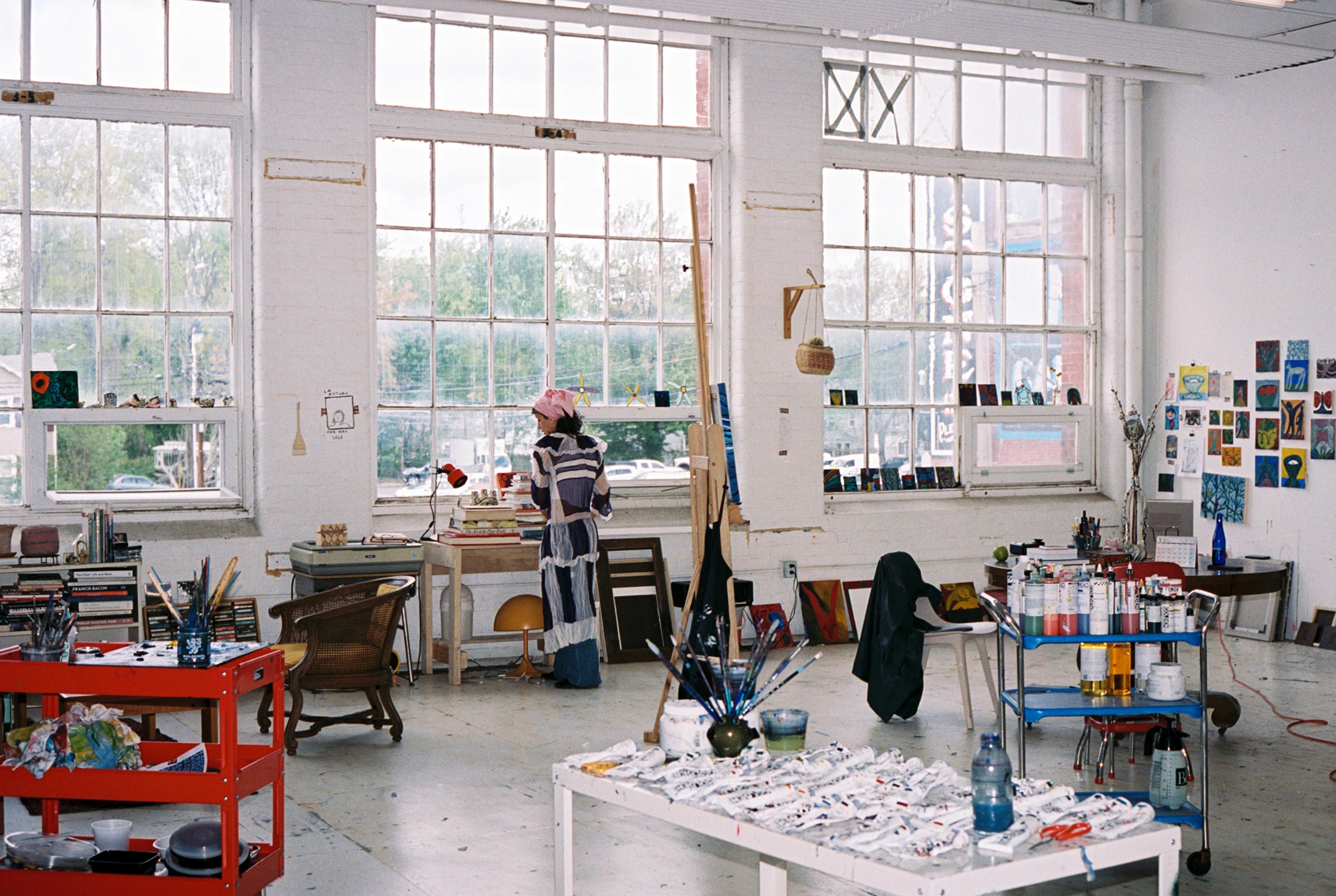 Artist Emma Kohlmann standing centrally in her studio space which has white walls, three large windows and trollies covered in paints and inks