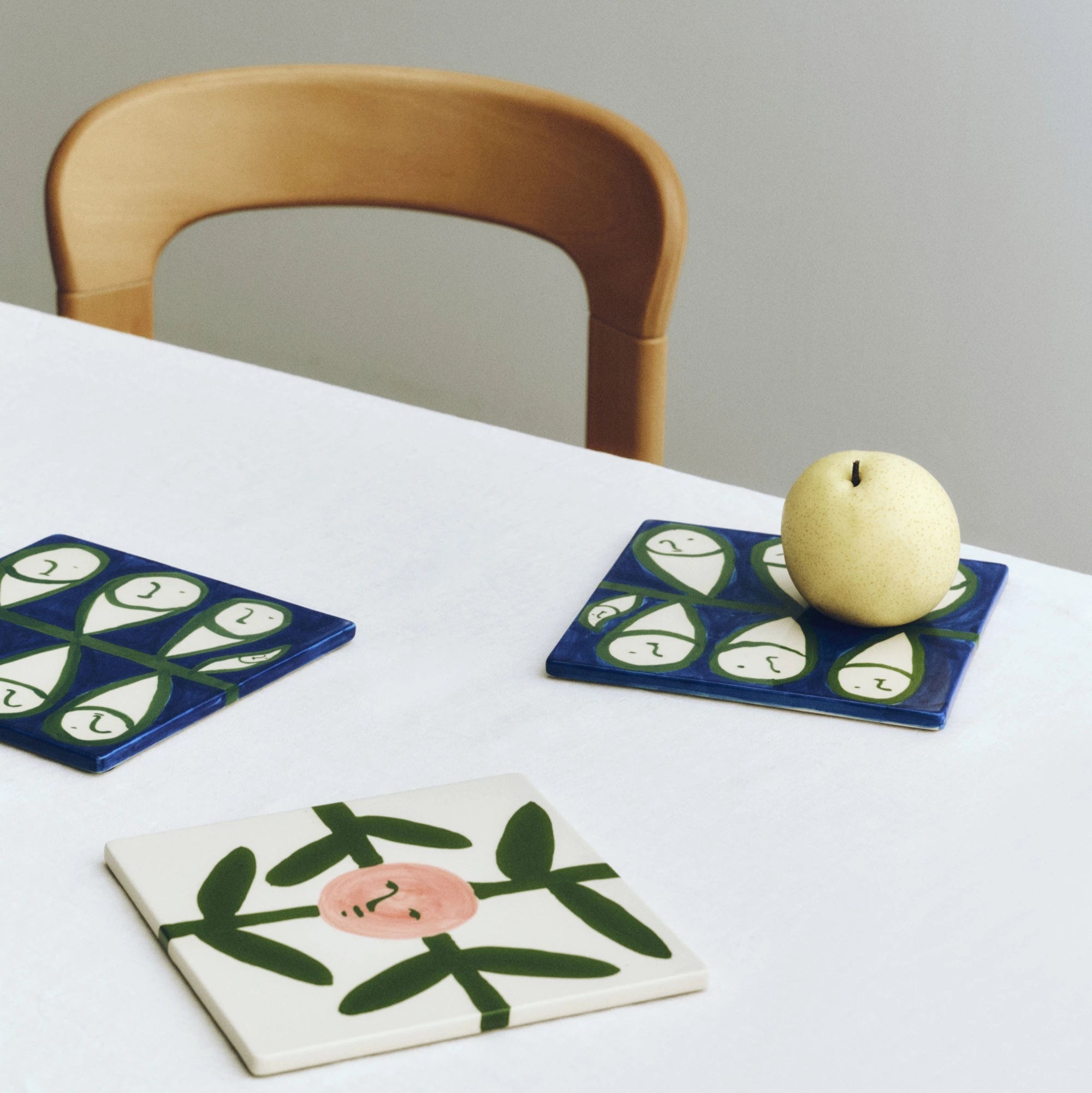 HAY x Emma Kohlmann - La Pittura Trivets on a white table clothed surface, one has an apple on it and in the background is the back of a wooden chair