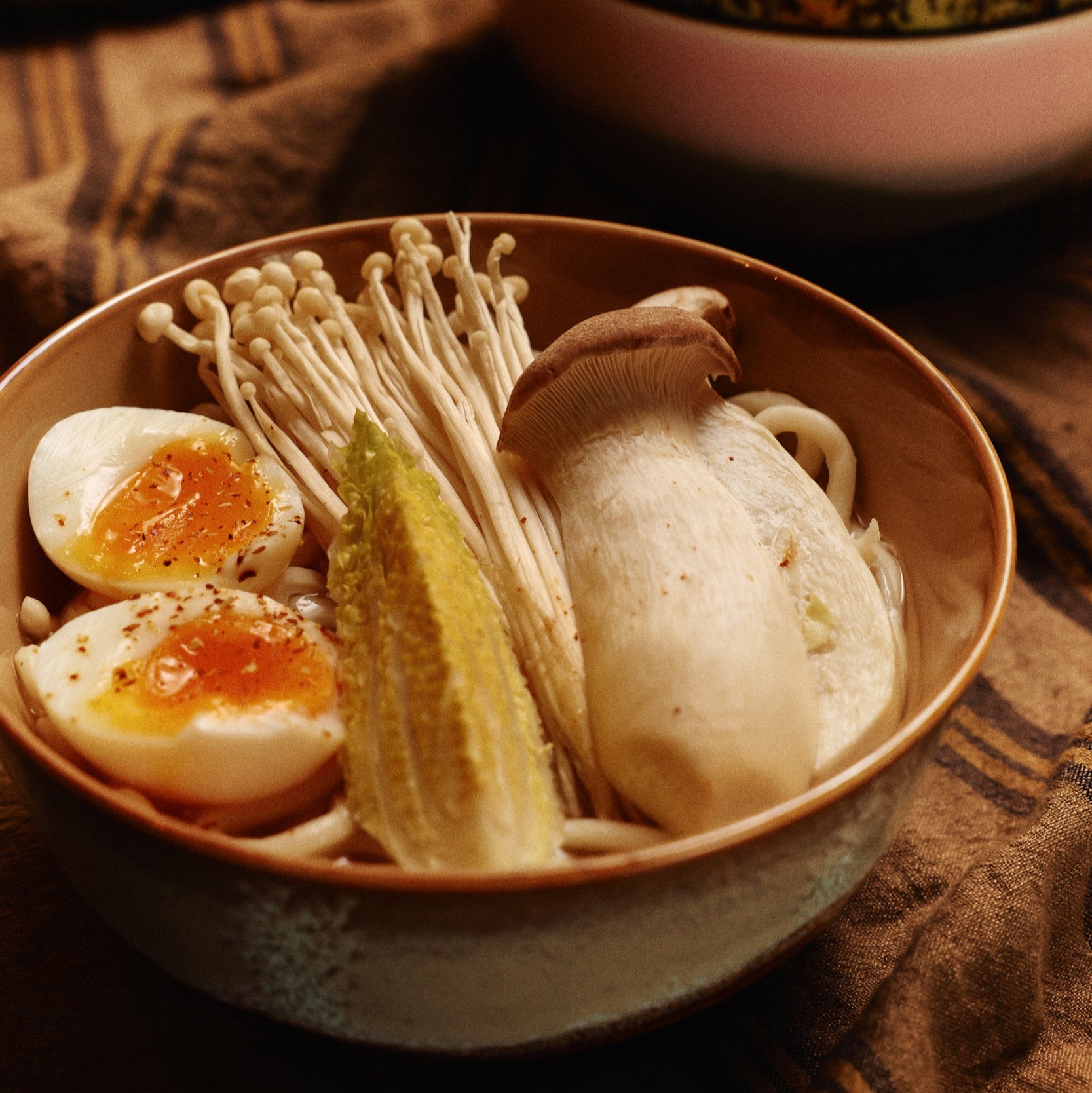 Bowl of Japanese cuisine with enoki mushrooms, pickled radish, and boiled eggs on a textured fabric background.