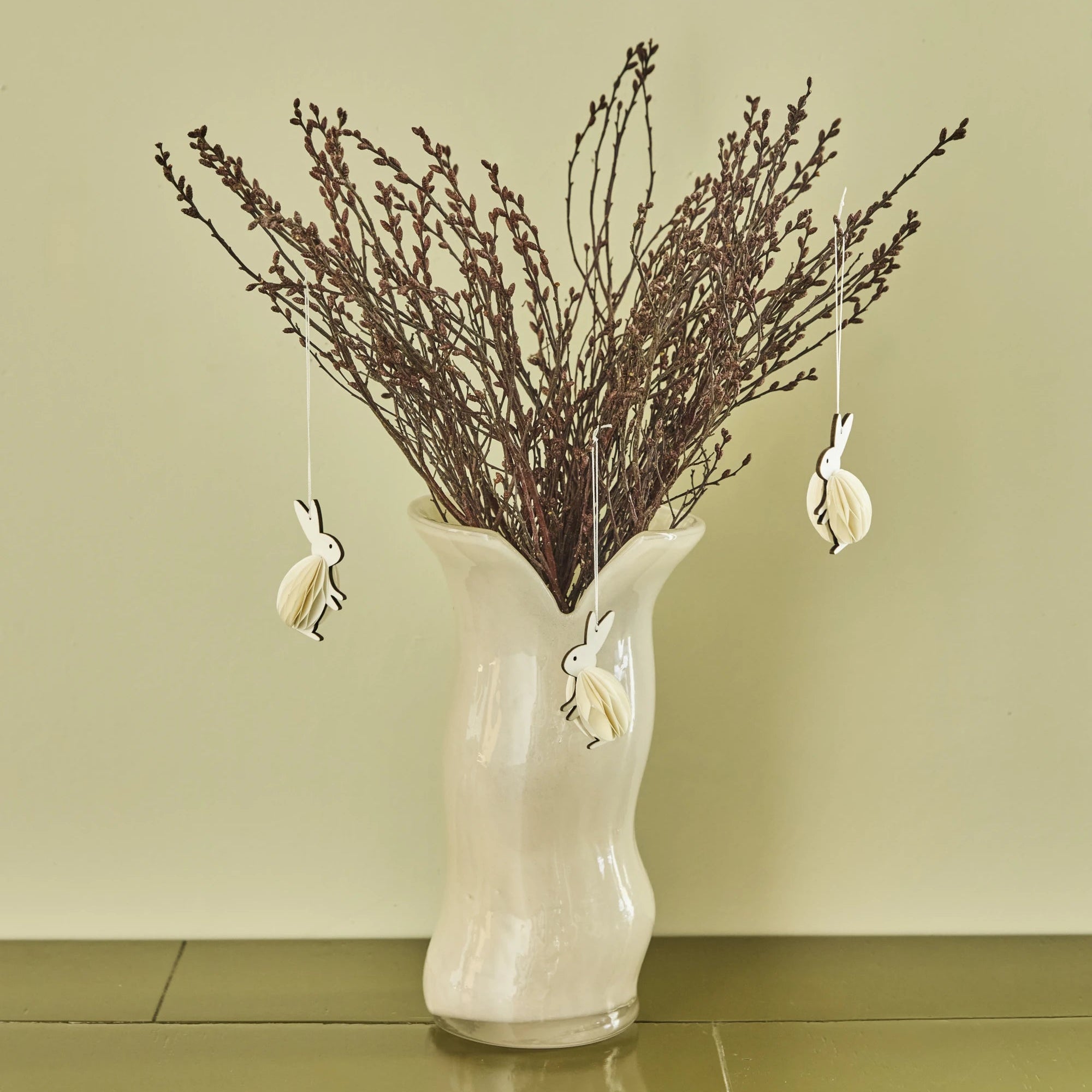 White vase with dried branches and hanging rabbit ornaments against a light green wall