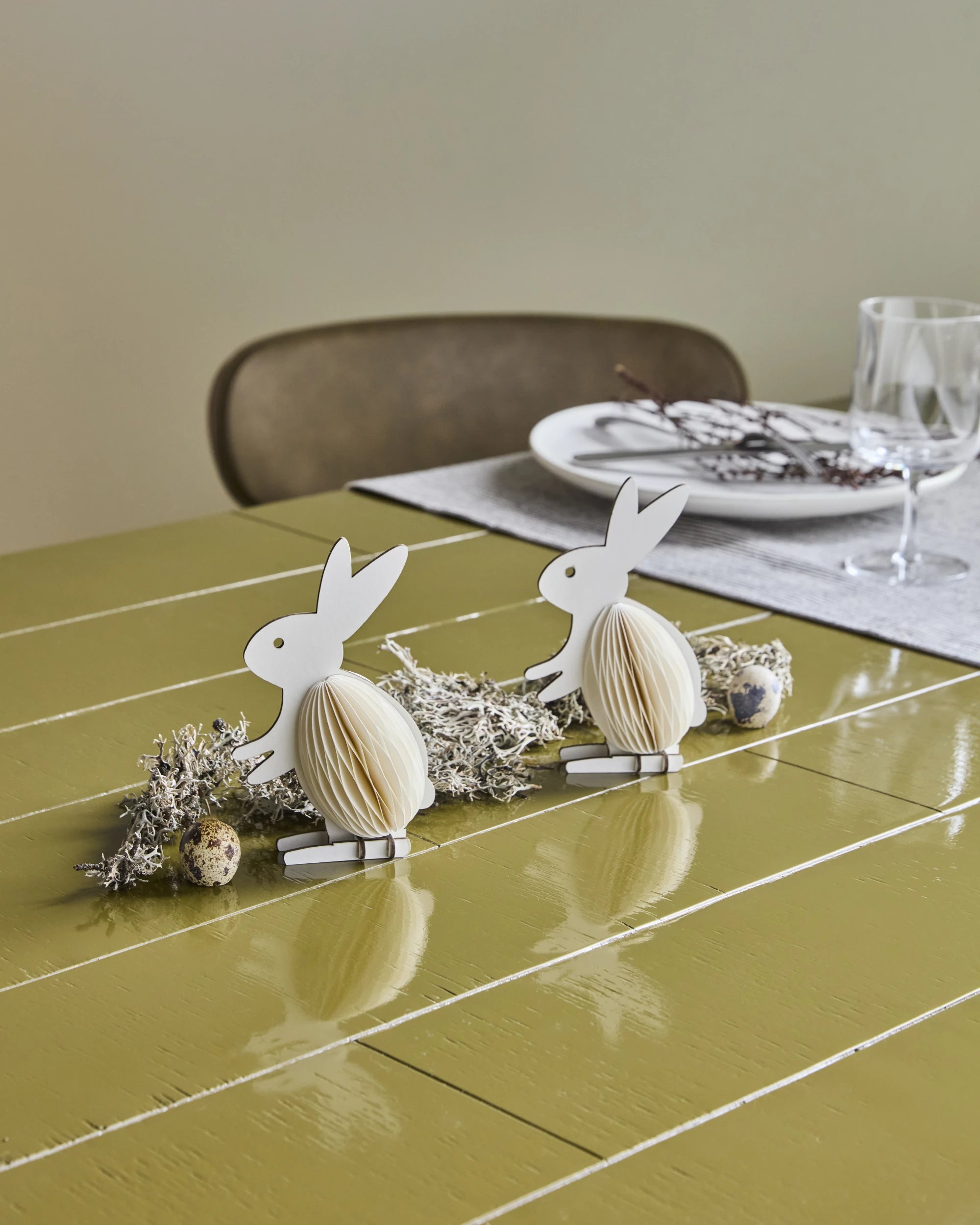 a green table with a place setting and 2 white paper standing rabbit ornaments next to lichen and eggs
