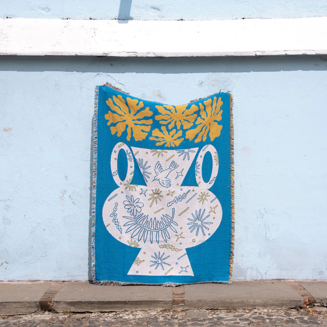 Blue, white and yellow tapestry blanket depicting a white vase and yellow flowers against a light blue wall