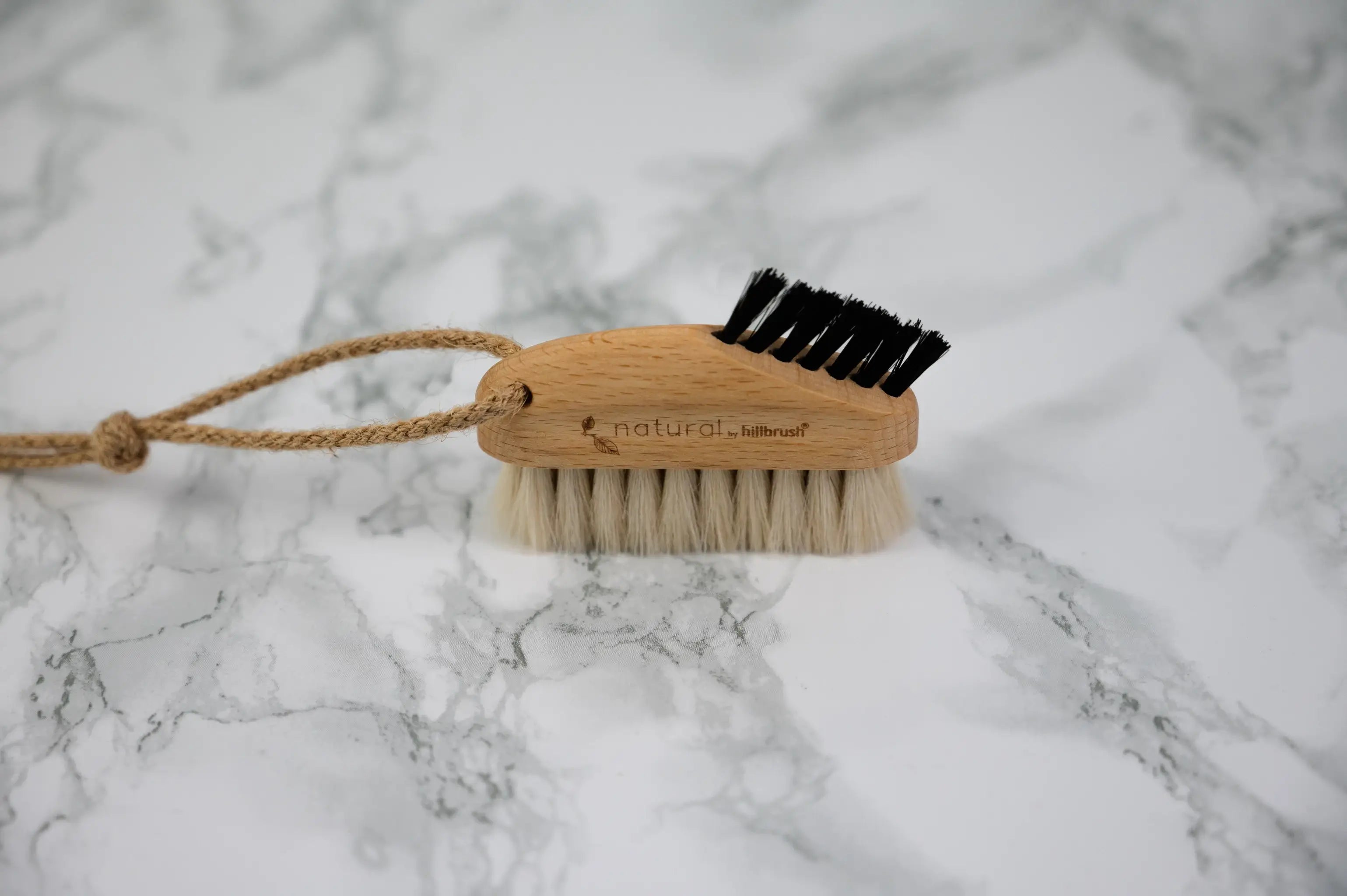 A small wooden laptop brush with two different sections of bristles and a hanging loop sits on a marble surface. Made by UK makers Hillbrush