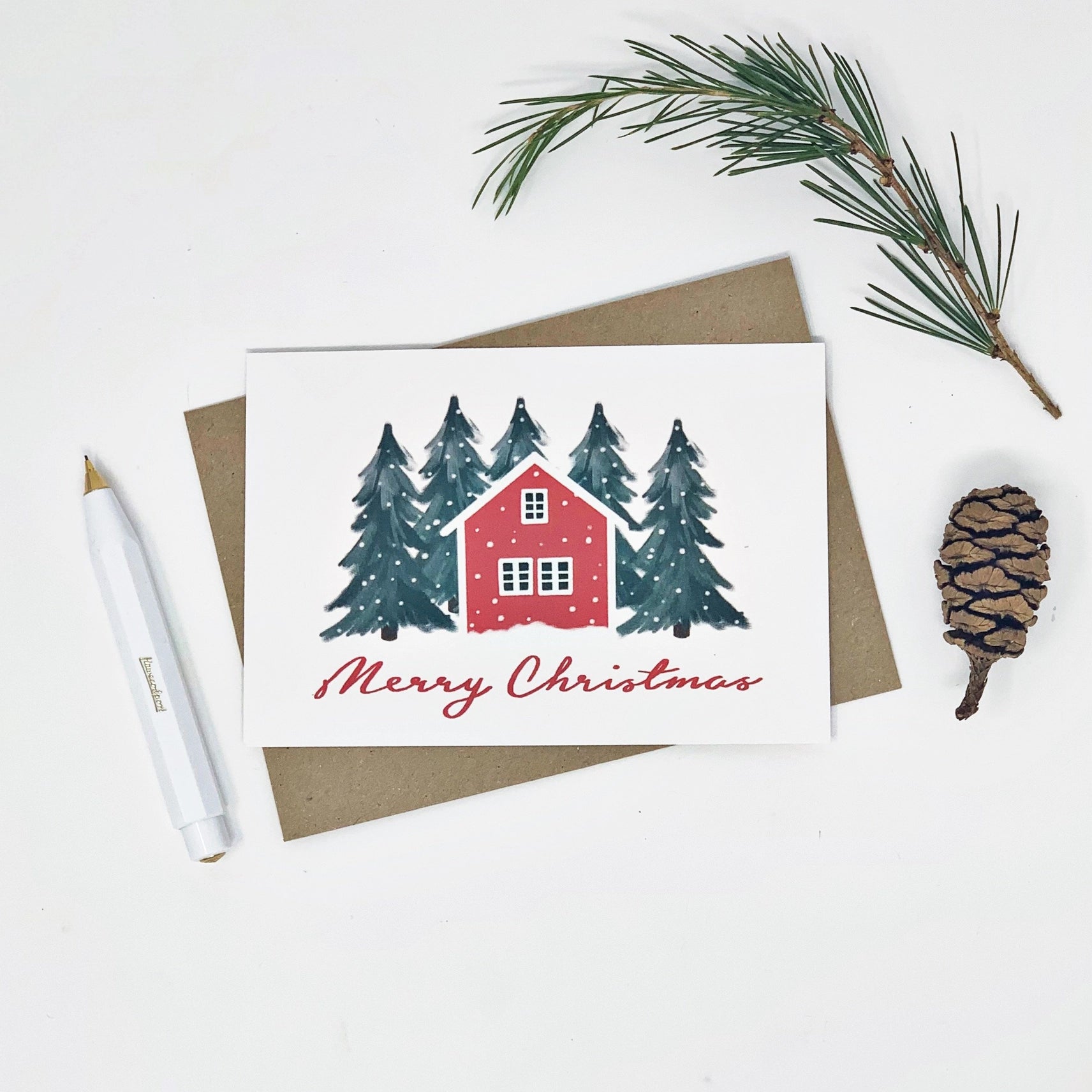 Christmas card with a red house and trees, surrounded by pine branches and a pine cone on a white background.