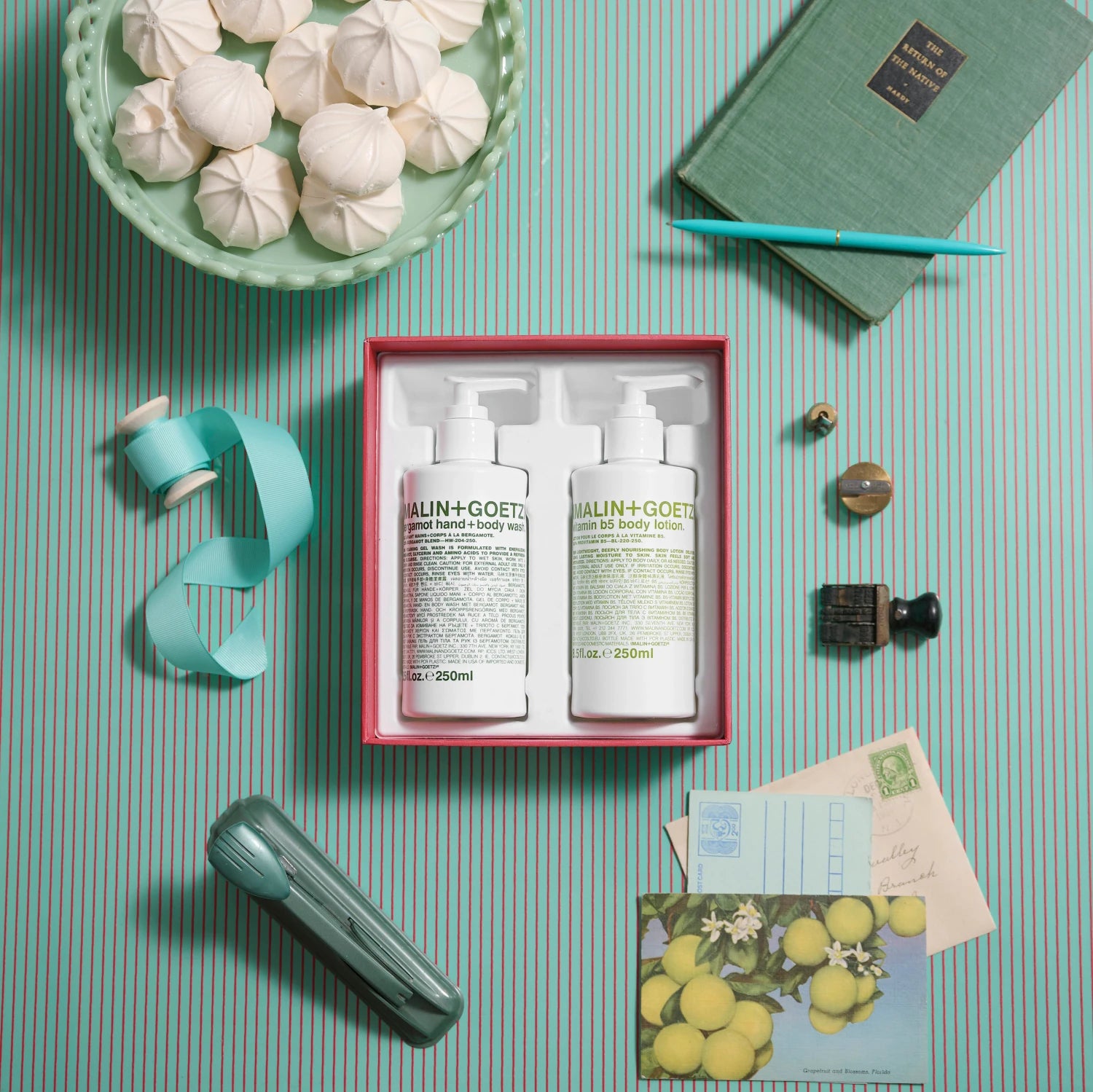 Malin and Goetz The Bright Side Bergamot Hand and Body gift set as a flatlay image on a teal and red striped background surrounded by teal stationery