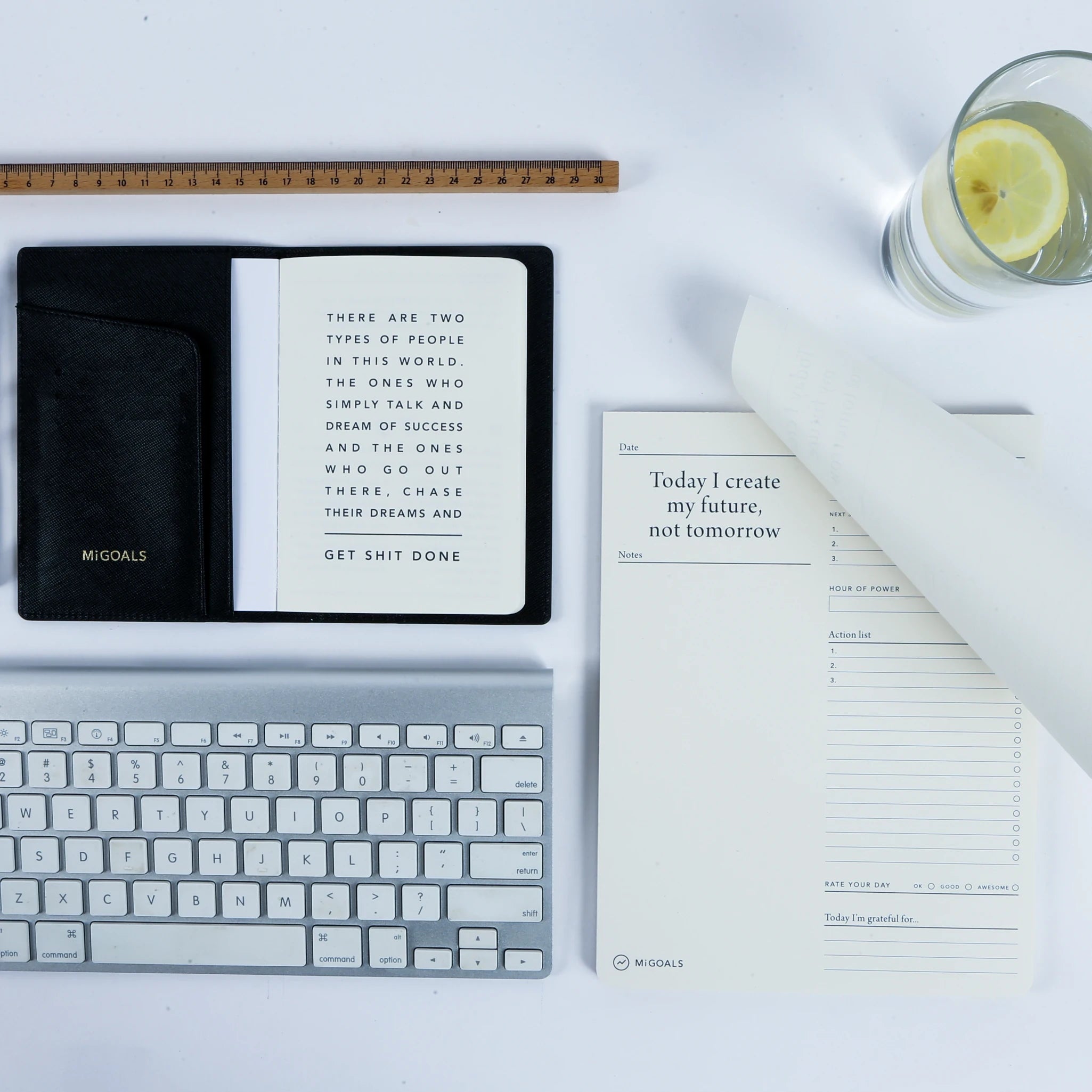 A flatlay image of a white background with the MiGoals To Do Desk pad, a keyboard, a drink, a ruler and another notebook