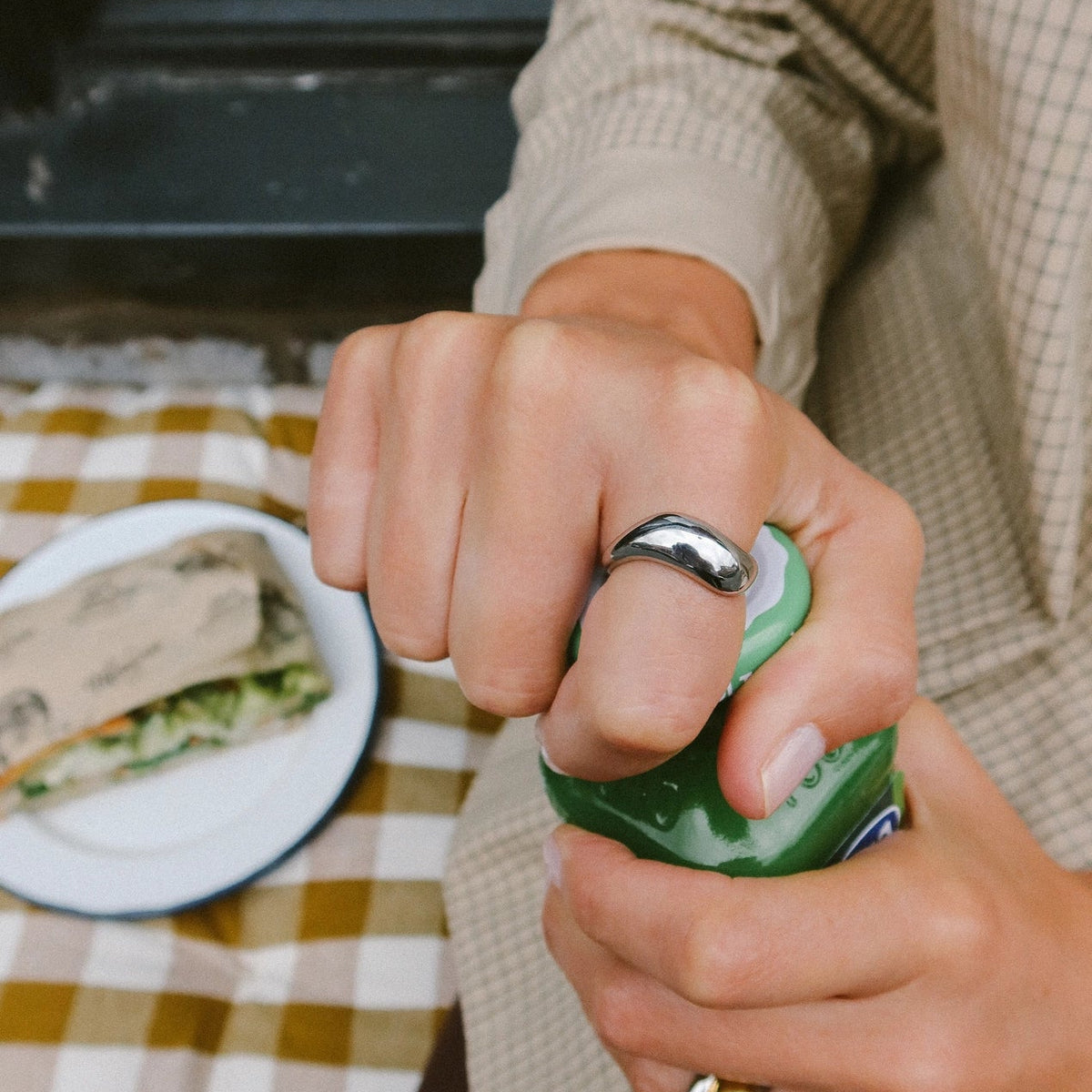 A close-up lifestyle shot of a person's hands wearing silver rings, one of which is being used to open a green can. The person is wearing a beige checkered shirt, and a plate with a sandwich is visible in the background on a yellow-and-white checkered tablecloth.