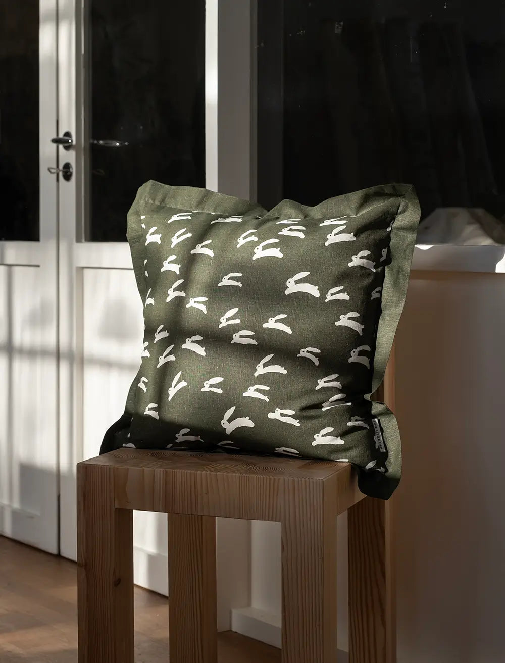 On a wooden chair sits a square moss green cushion with playful white rabbit print by Fine Little Day