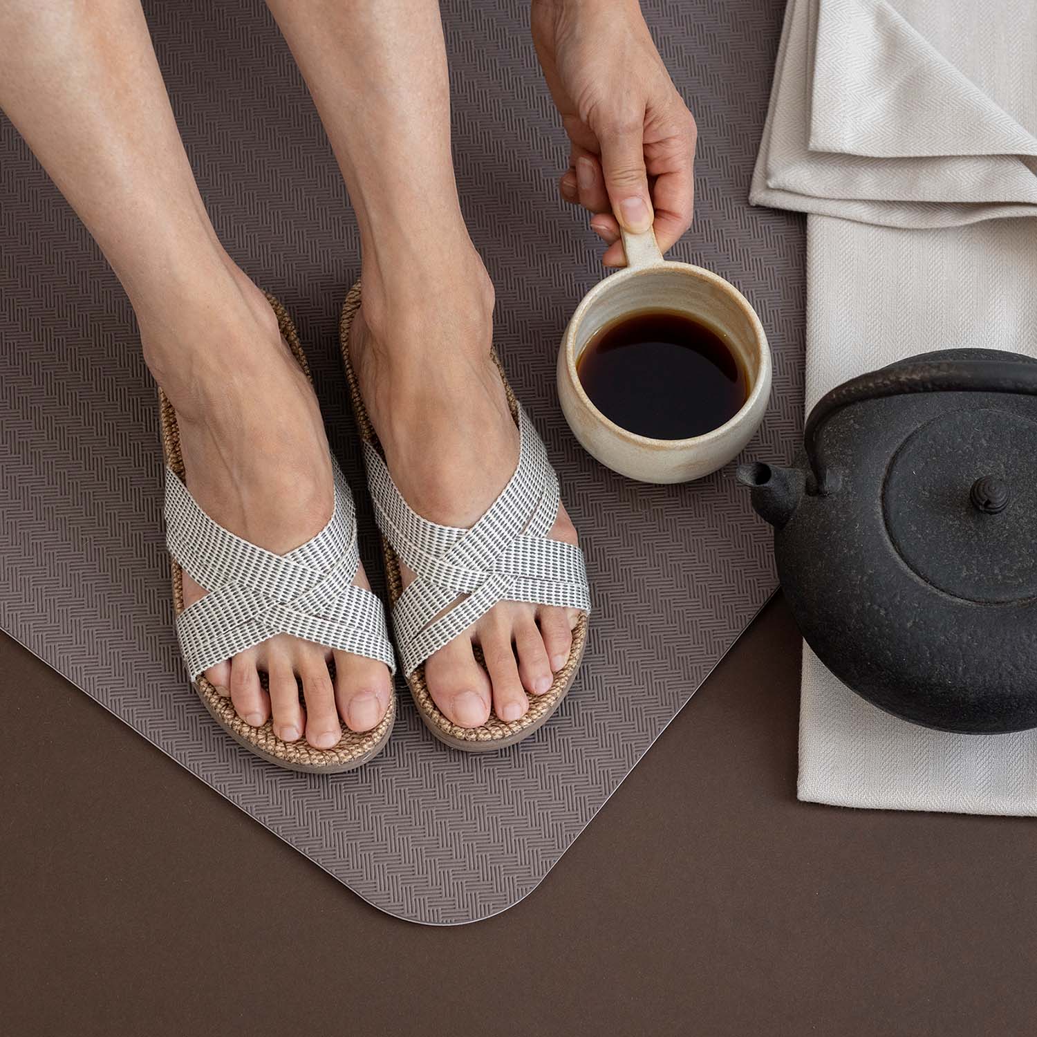 A pair of womens feet wearing Shangies sandals on a yoga mat next to a teapot and full mug of black tea