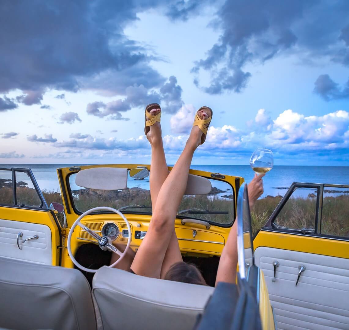 A woman lies back in her stationary open topped yellow car and balances her legs on the edge of the frame whilst wearing yellow Shangies sandals