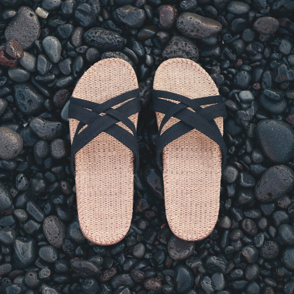 A top view of a pair of womens Shangies sandals in black and natural jute on a background of smooth black stones