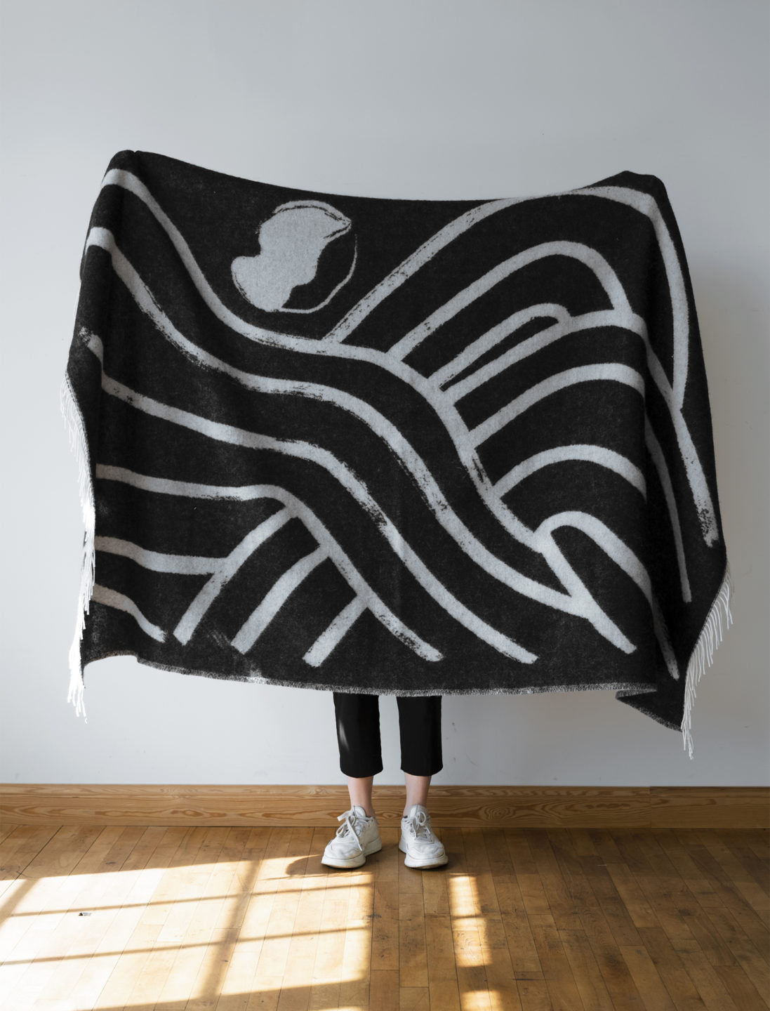 A person folds a full size dark grey with light grey patterned lambswool blanket from a collaboration between Sofia Lind and Fine Little Day