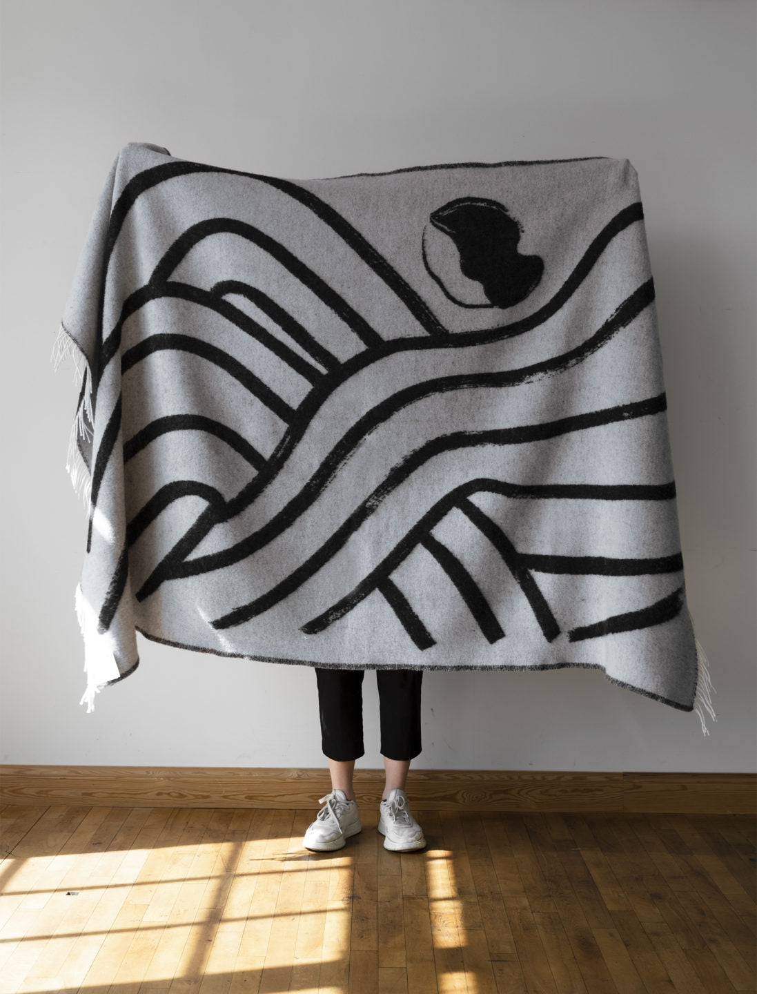 A person folds a full size light grey with dark grey patterned lambswool blanket from a collaboration between Sofia Lind and Fine Little Day