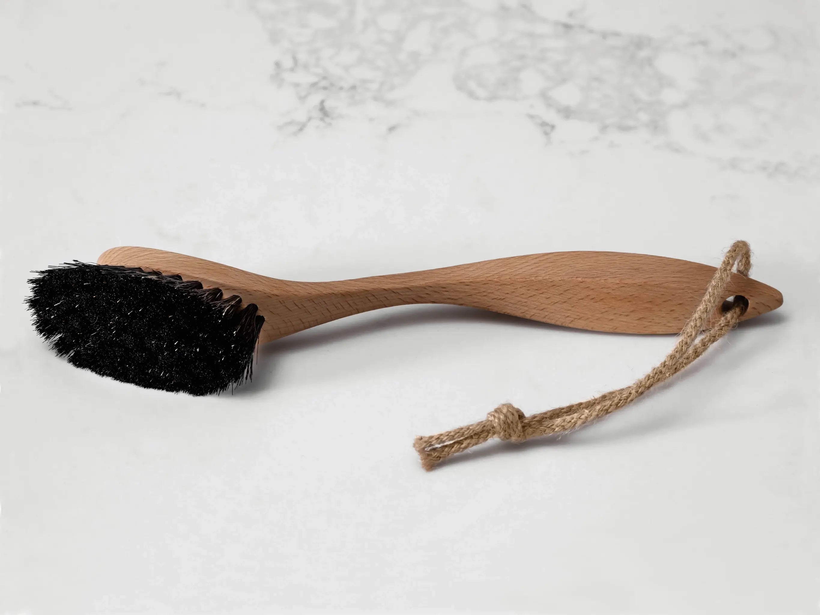 A long handled wooden dish brush with dark bristles and a hanging loop sit on a marbled surface. Made by UK makers Hillbrush