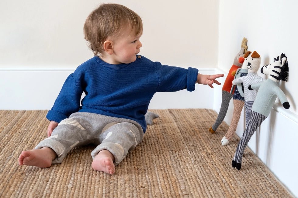 Baby in a dark blue jumper reaching towards several Sophie Home stuffed toy animals