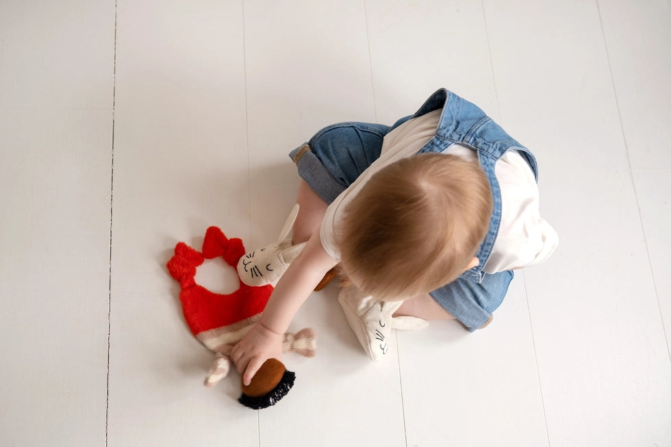 Red Buddy Comforter Cuddle Cloth by Sophie Home on a white floor next to a baby in blue overalls