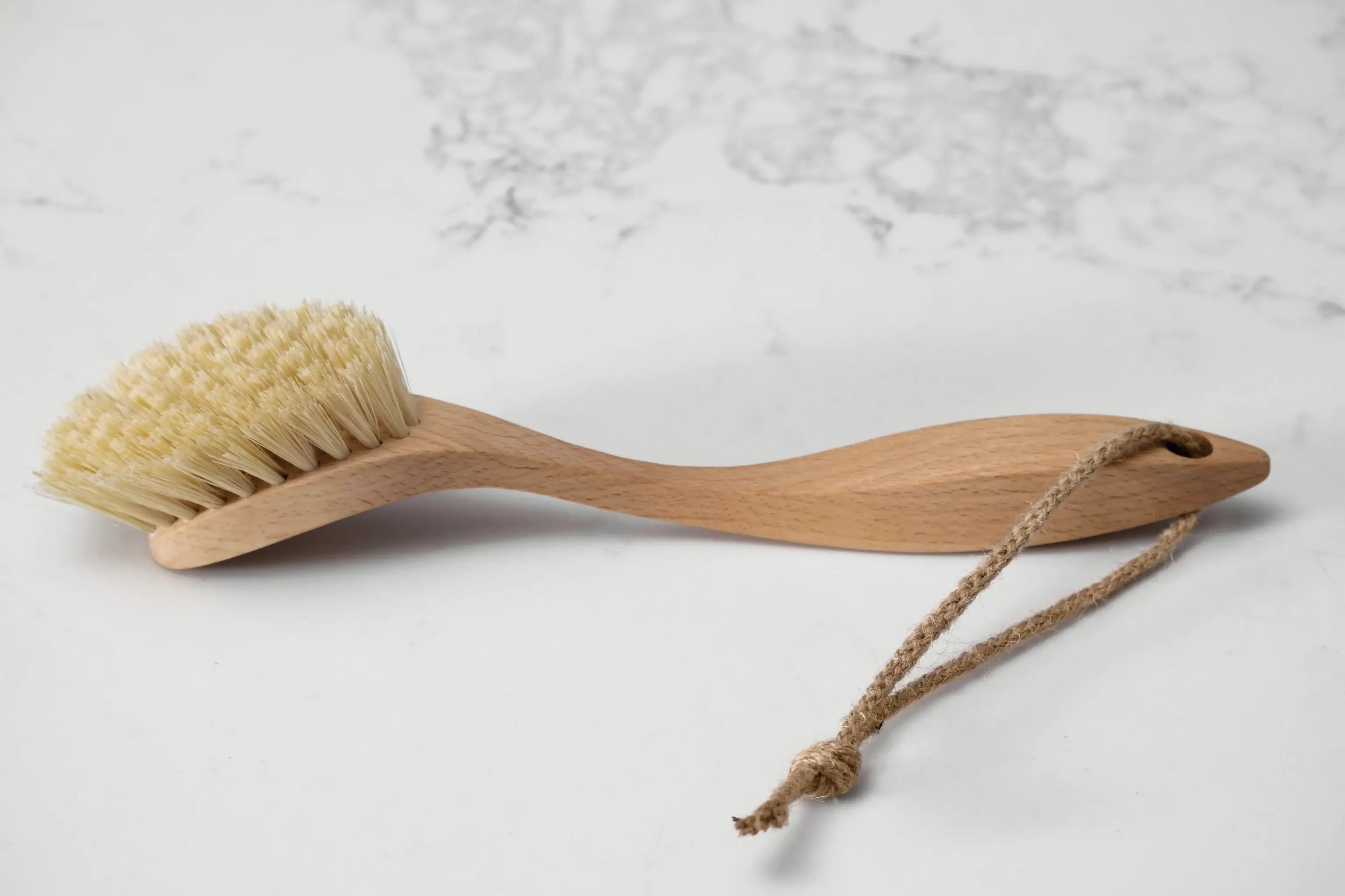 A long handled wooden dish brush with thick light coloured fibres and a hanging loop sit on a marbled surface. Made by UK makers Hillbrush