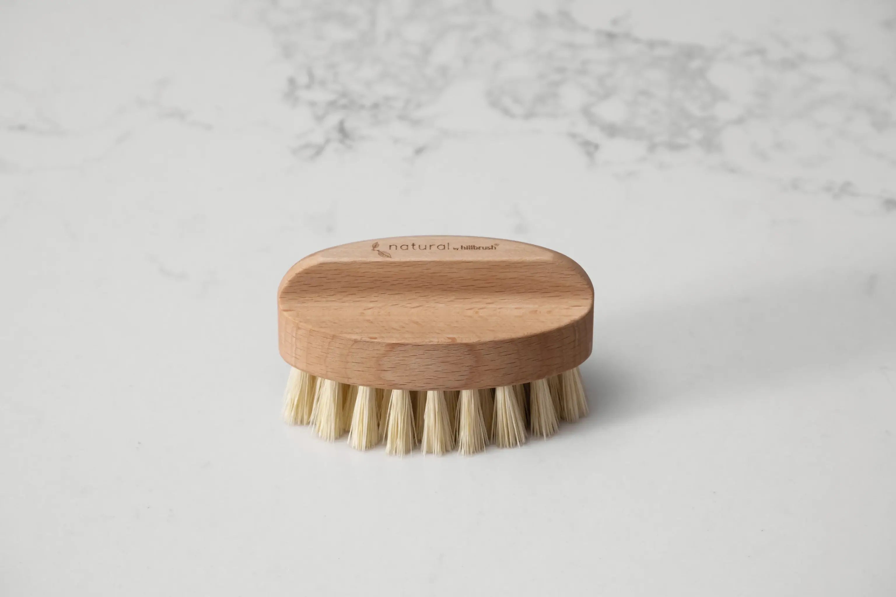 A small oval wooden nailbrush with thick fibres sits on a marbled background. Made by UK makers Hillbrush