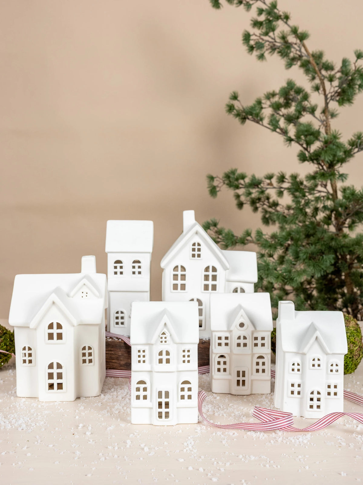 A set of small white ceramic Christmas Byn house ornaments by Storefactory with cutout windows, arranged on a light surface with a red-and-white striped ribbon and a blurred evergreen branch in the background.