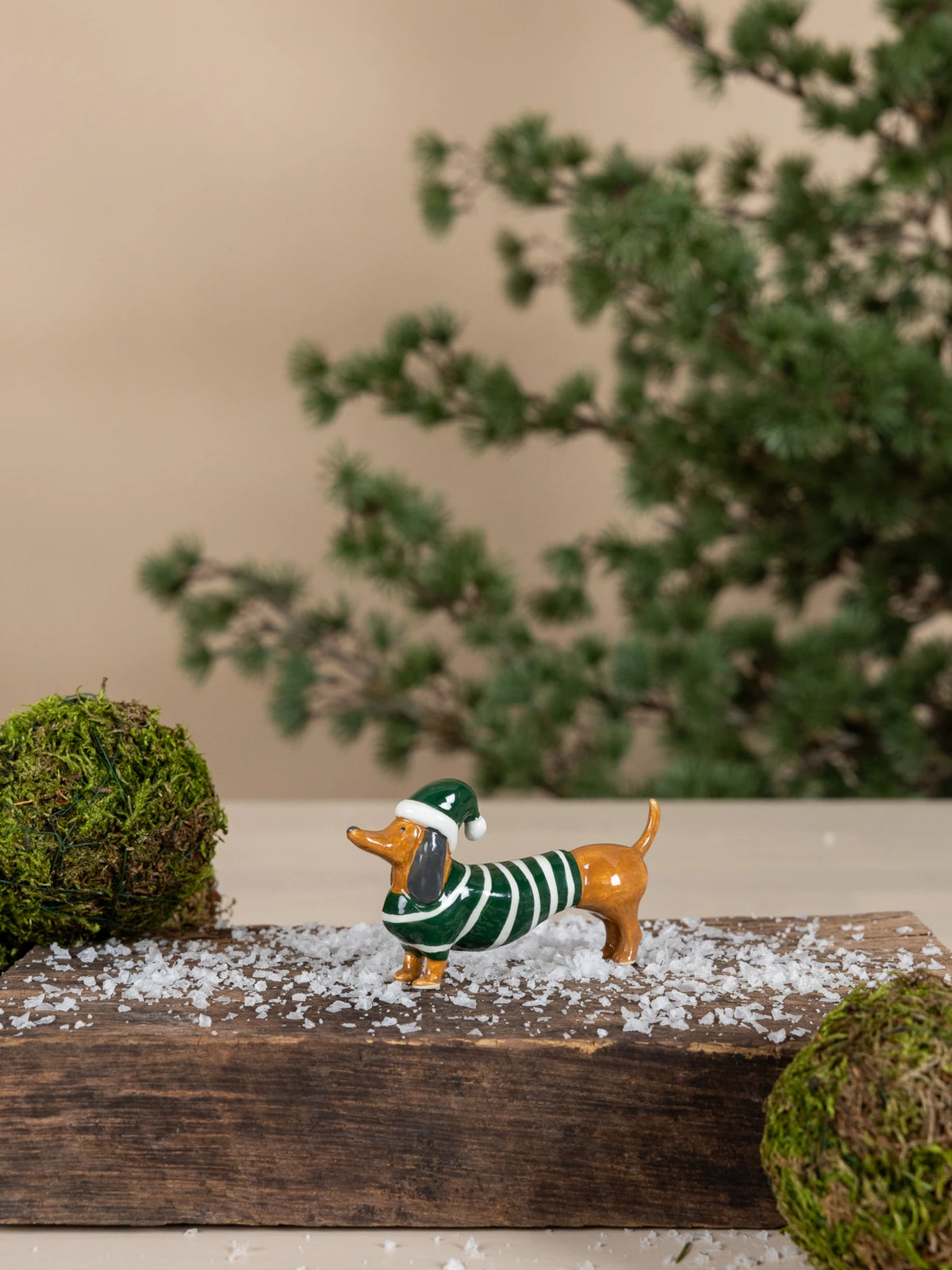 Small dog figurine wearing a green sweater called Albin by Storefactory on a wooden surface with a blurred plant in the background