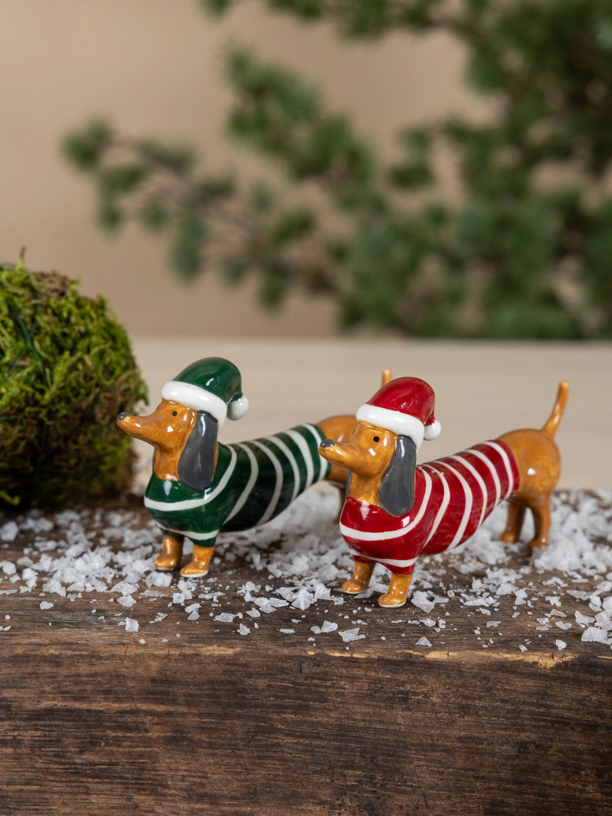 Two small dachshund Albin figurines by Storefactory dressed in holiday sweaters and Santa hats, standing on a wooden base with faux snow, moss balls, and a blurred evergreen background.