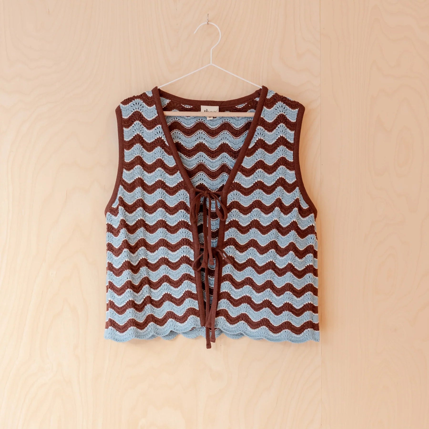 A sleeveless knit vest with a brown and light blue wavy chevron pattern, featuring a front tie closure, hanging on a white hanger against a light wood-paneled wall.