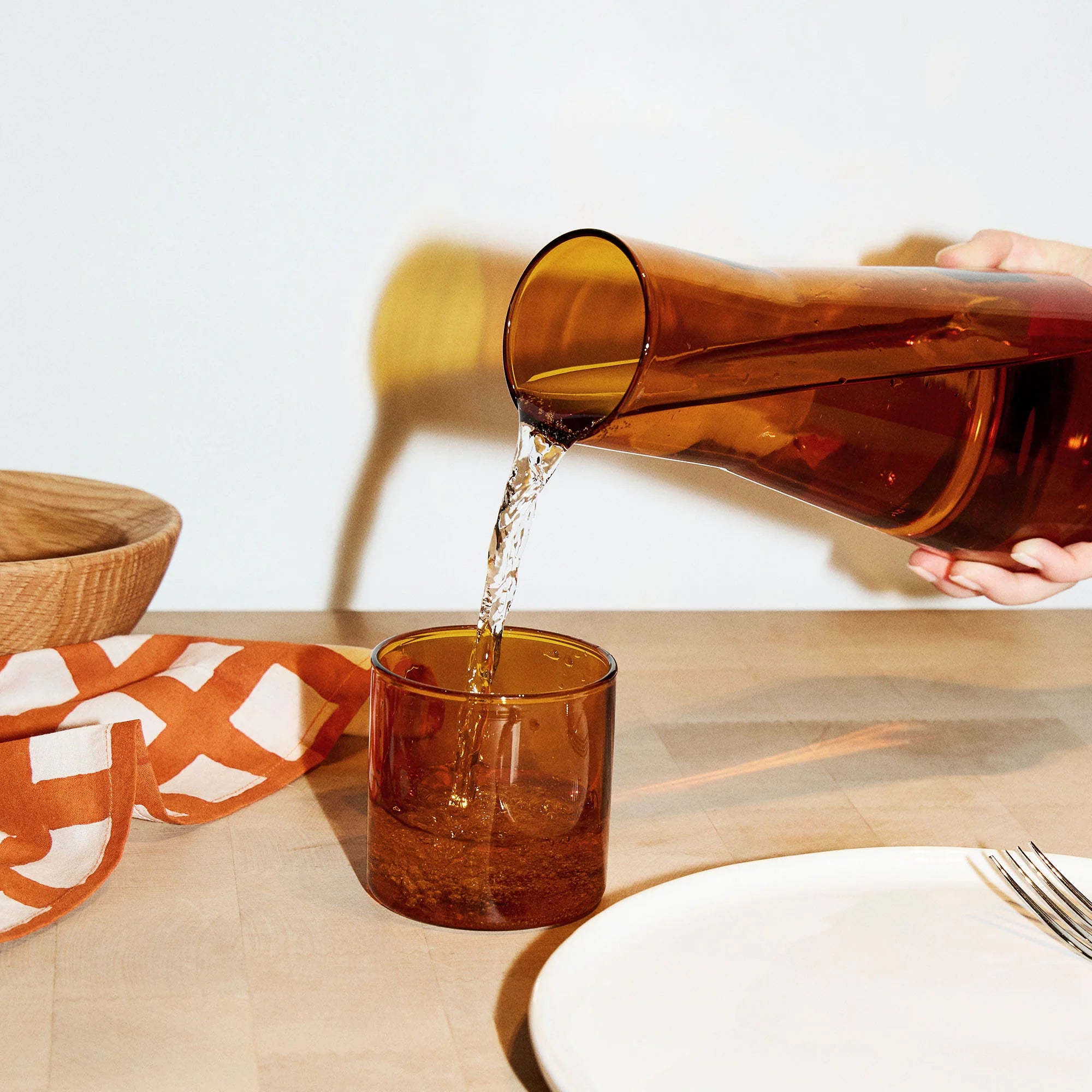 Hand holding an amber glass carafe pouring liquid into an amber glass on a wooden table with a white background
