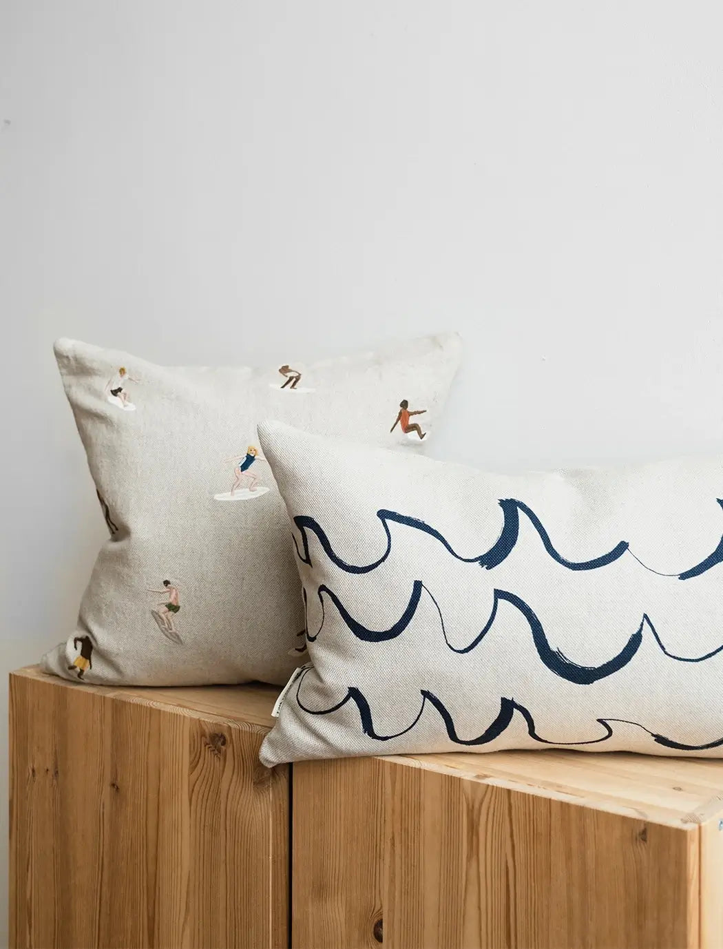 A long cushion in natural linen with a minimal blue wave shaped brush stroke motif from Fine Little Day. It sits next to a square cushion with embroidered surfers on it.