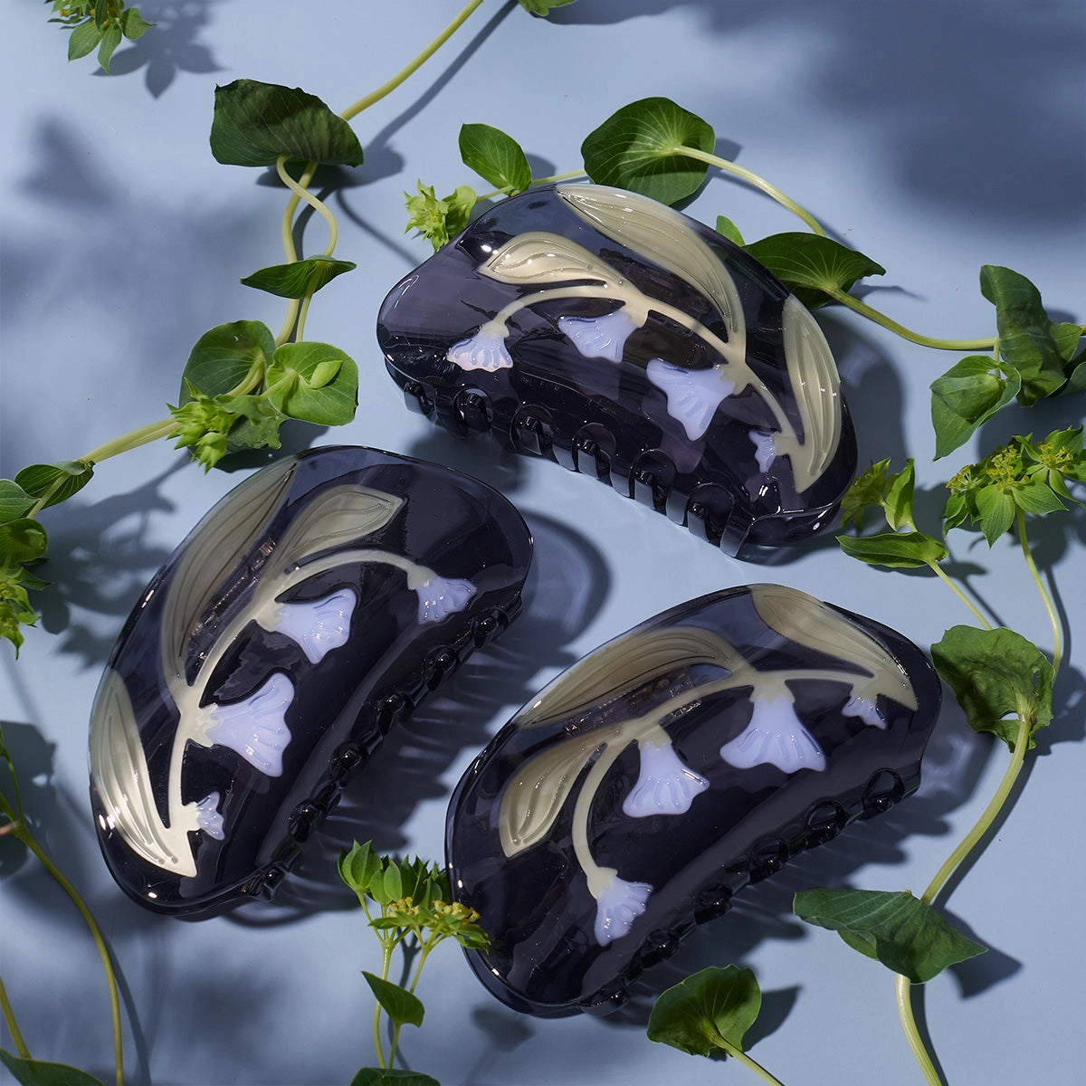 Three black hair clips with white floral designs on a light blue background with green leaves.