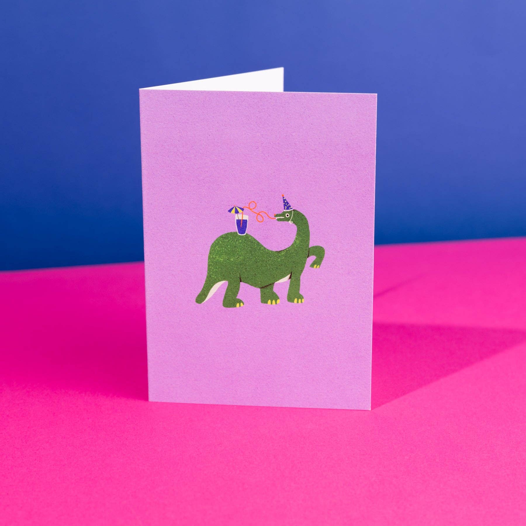 Standing card with a green dinosaur wearing a party hat design on a purple background, against a blue and bright pink background