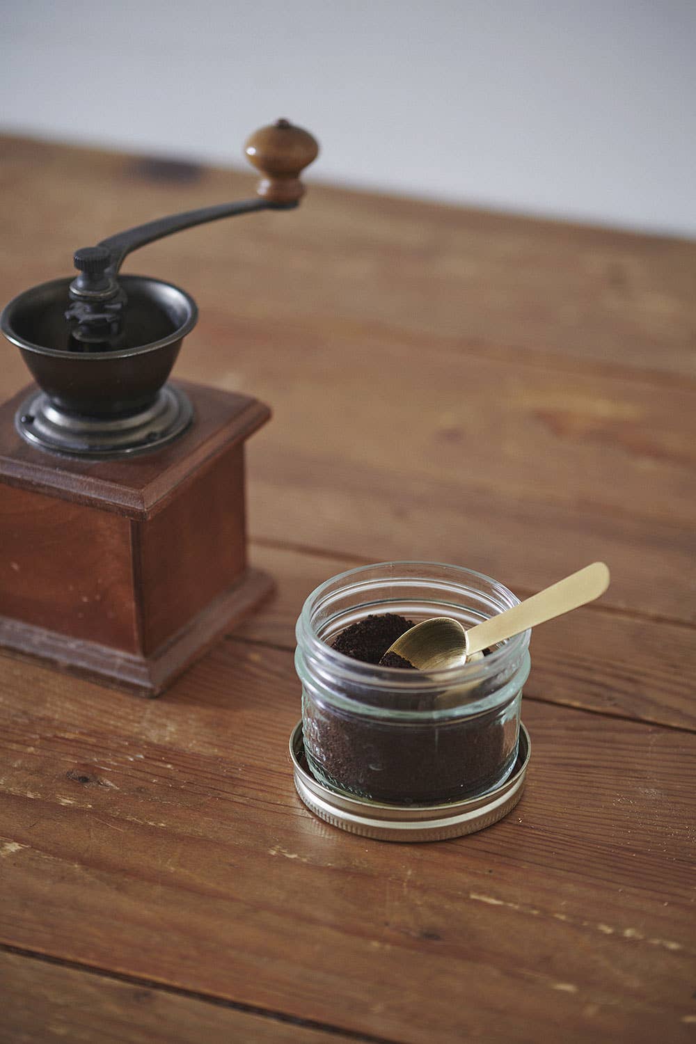 Jar of coffee grounds with a spoon next to an old-fashioned coffee grinder on a wooden surface.