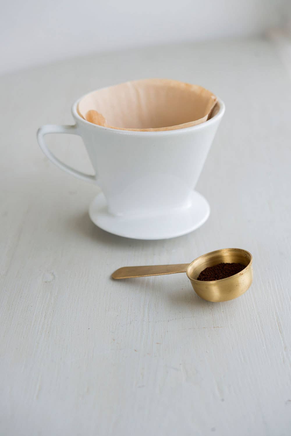 White coffee filter in a V60 with a brass scoop of coffee grounds on a light surface