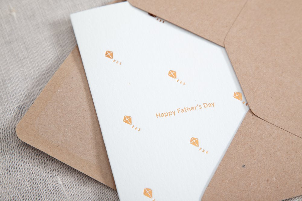 A natural white card with copper writing saying Happy Fathers Day, surrounded by tiny kite images. The card is coming out of a brown envelope.