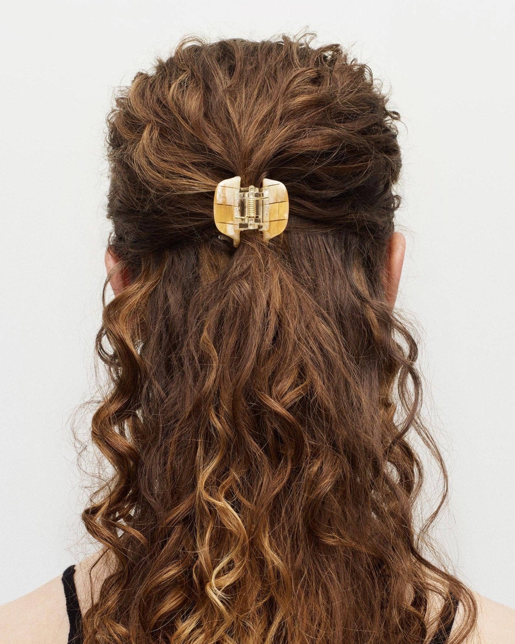 The back view of a white female model with brown curly long hair wearing the MACHETE mini hair claw with her hair in a half up style
